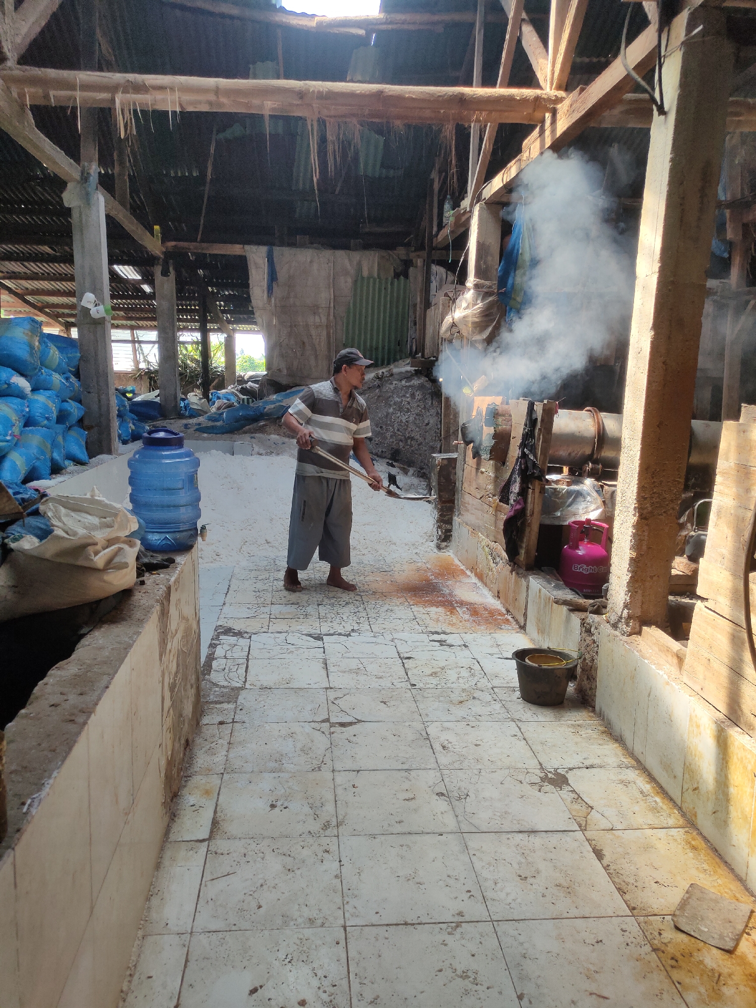 A man wearing a striped shirt, shorts, and a baseball cap is working in a rustic, open-air structure. He is holding a long-handled tool and appears to be stirring or raking a substance on the tiled floor. White smoke billows from equipment to his right, suggesting some form of processing or cooking is taking place. Large blue sacks are stacked to the left of the man, and a large blue water cooler is positioned near them. A pink propane tank is visible near the smoking equipment. The building has a corrugated roof and wooden beams. The floor is tiled but appears worn and dirty. The scene is outdoors, with sunlight filtering through gaps in the roof, indicating it is daytime. This scene is located in Sumedang, Indonesia, a region known for its agricultural processing and local industries.
