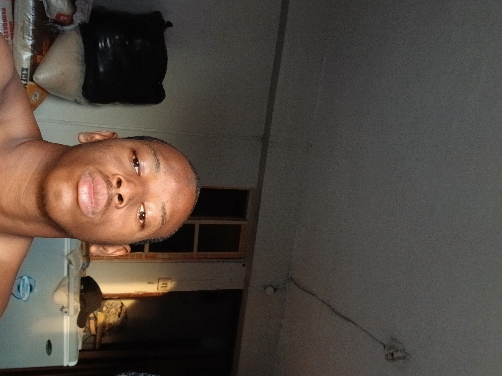 A close-up, chest-up selfie shows a young Black male, shirtless, looking directly at the camera with a neutral expression. He has a close-shaven head and full lips. The image is oriented vertically, with the subject positioned on the left side of the frame.
The background indicates an indoor setting, likely a room or kitchen, characterized by dim lighting. To the upper left, several bags are stacked; one clear plastic bag contains white grains and is labeled "PARBOILED INDIAN". Another bag is dark-colored. Below these, a portion of a white refrigerator or large appliance is visible, featuring a sticker with "PREMIUM" text.
In the mid-ground behind the subject, a window frame with dark panes suggests the scene is dimly lit or it is dark outside. Below the window, a dark hat is placed on a surface. The ceiling and upper wall on the right are plain and light grey, with visible electrical wires and a small, round object (possibly a sensor or switch) attached to the wall. The overall scene suggests a candid moment in a domestic environment in Lagos, Nigeria.
