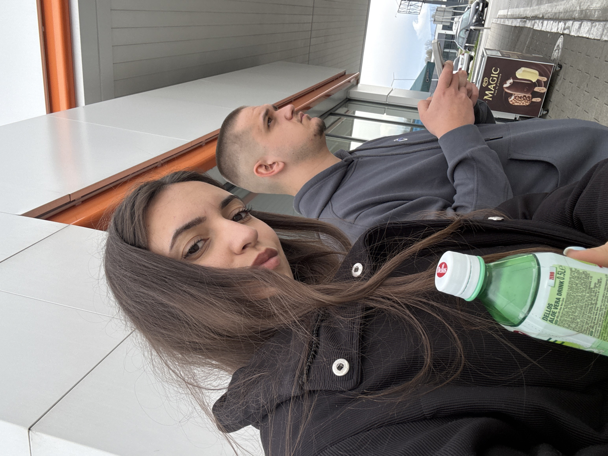 A woman with long brown hair and a black jacket holds a bottle of "Dellos Aloe Vera Drink 0.5L," posing for a selfie. Behind her is a man with short dark hair wearing a gray hoodie, looking away while holding a phone. An advertising stand for "Magic" ice cream is visible in the background. The location appears to be outdoors near a modern building, possibly in a parking area of a shopping center, in the city of Unknown, Bulgaria.