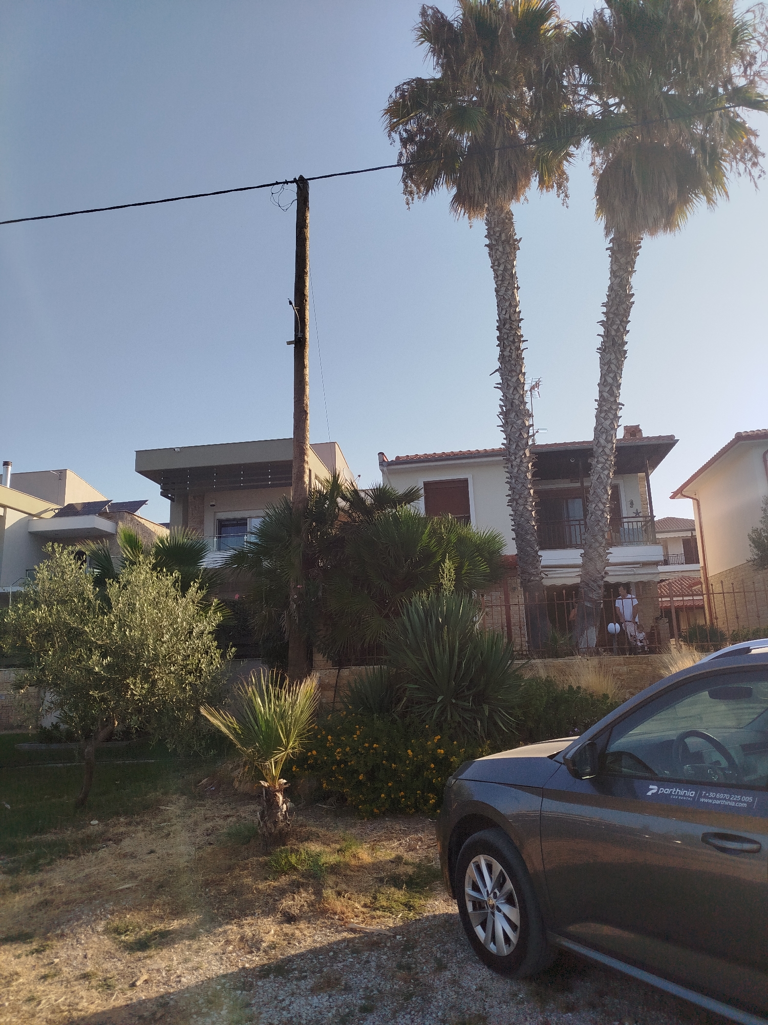 The image captures an outdoor scene on a clear day with a bright sky, likely in Sithonia Municipal Unit, Greece. In the foreground, the right side of a dark gray car is visible. The car has a sticker on its side with the text "parthinina CAR RENTAL" and a phone number and website. Beyond the car, the ground is covered with gravel and dry grass.
A fence made of stone and metal bars separates the foreground from a property with vegetation and buildings. A prominent feature in the background is a large wooden utility pole with wires running across it. To the left of the pole, there are several buildings, including a modern house with solar panels on its roof. Lush green trees, including an olive tree, and some spiky plants are present on the property. Two tall palm trees stand in front of another building. This building appears to be a multi-story house with balconies and tiled roofs. A person is partially visible near the entrance of this house. The overall impression is a residential area with Mediterranean vegetation.