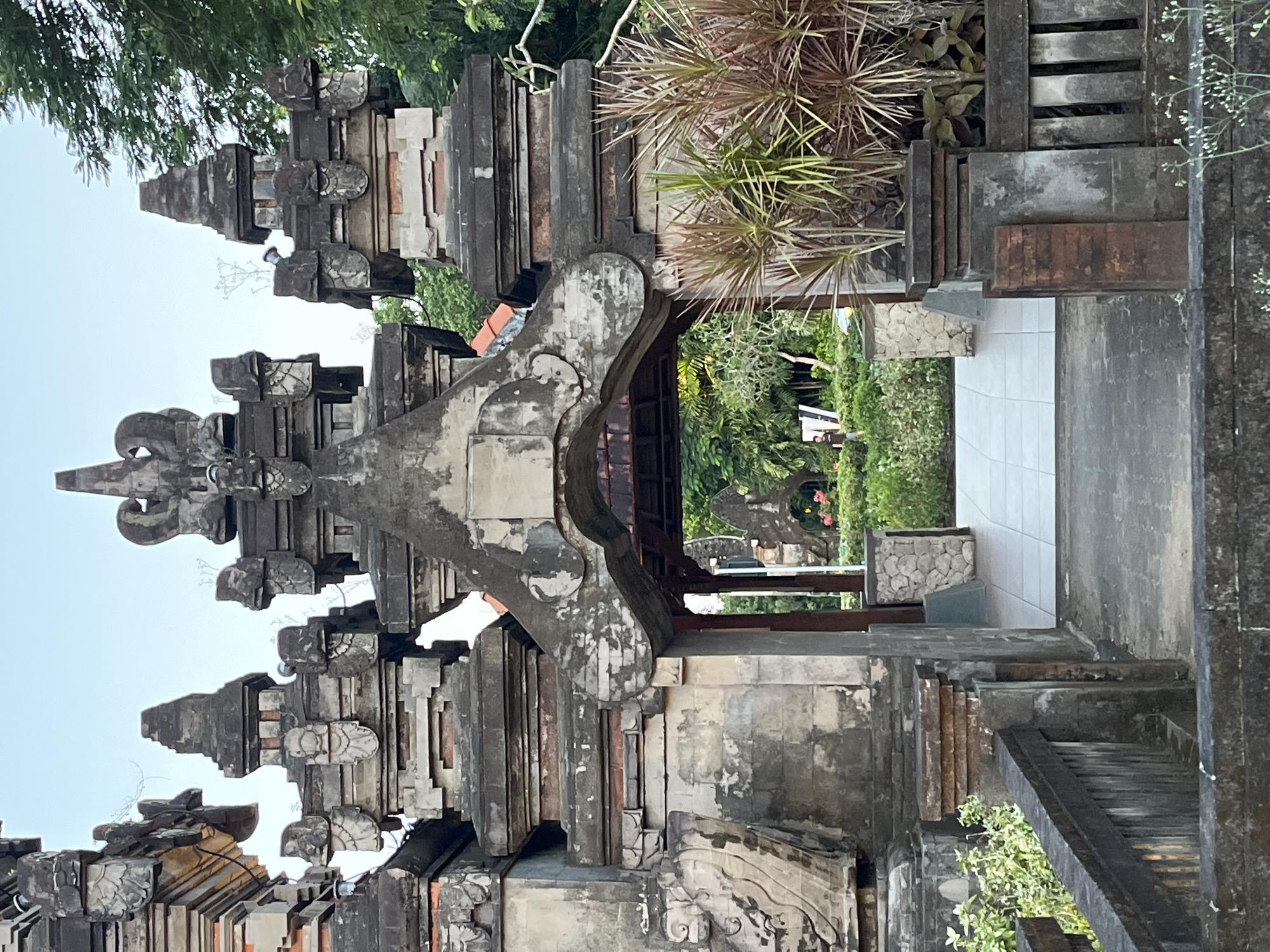 The image shows an ornate stone gateway, likely the entrance to a temple or significant structure, located in Unknown, Indonesia. The gateway is constructed from intricately carved, weathered stone and features a tiered roofline with decorative elements. The structure is gray and brown in color. Through the gateway, a glimpse of a garden or courtyard can be seen, with lush greenery and a tiled pathway leading deeper into the space. The sky is visible above the structure.