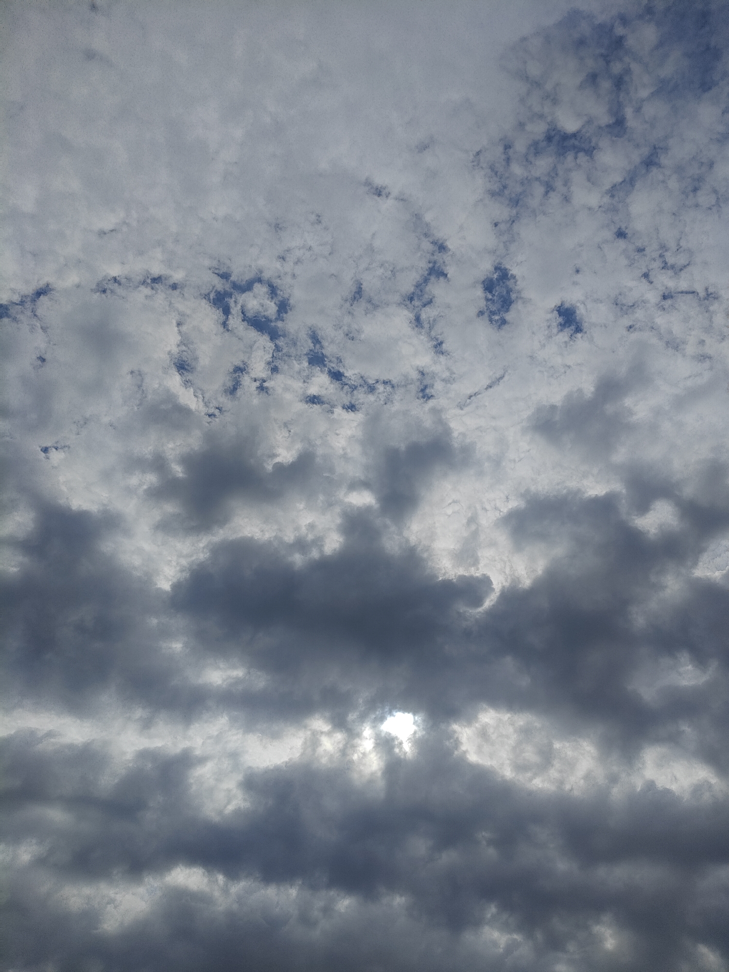 This is a medium shot, landscape orientation, photograph of a cloudy sky. The clouds are varied, with some appearing bright and fluffy, while others are dark and heavy. In the middle of the frame, a patch of blue sky is visible through a break in the clouds. The sun is peeking through a gap in the clouds, creating a bright spot in the image. The lighting suggests it is daytime, with the sun not directly overhead. The overall impression is of a dynamic sky, possibly before or during a change in weather. There are no people or objects visible in the frame, only the sky and clouds. No text is present.