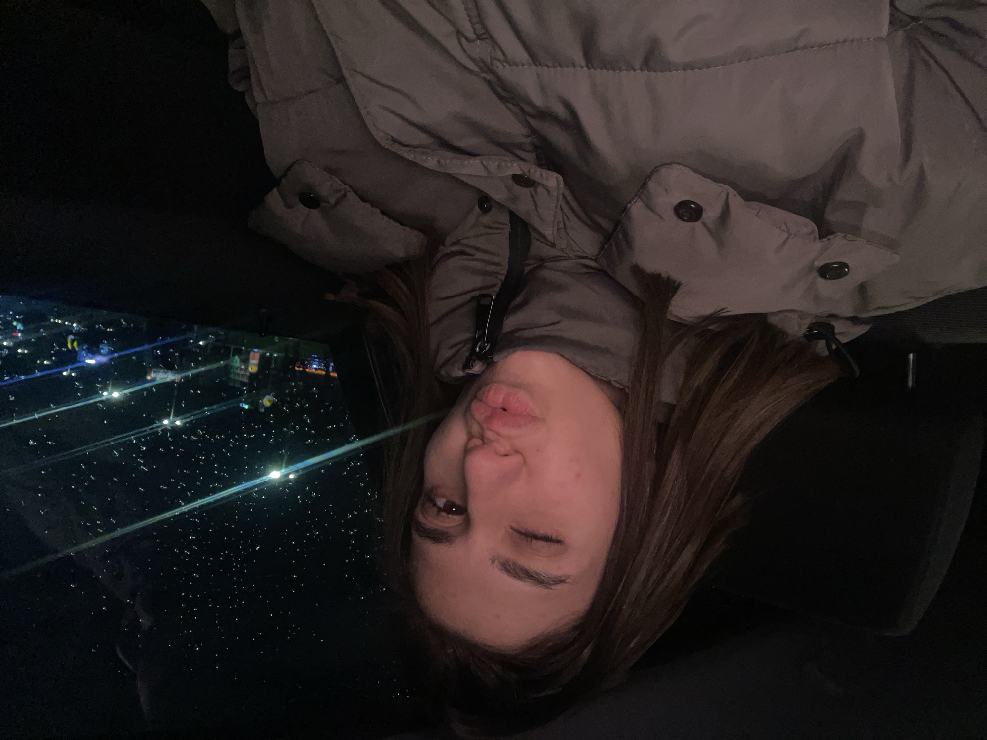 A young woman with light brown hair is sitting in what appears to be a car at night, making a kissy face and winking. She is wearing a puffy gray jacket. Through the car window, a nighttime view of a brightly lit urban area is visible, which might be identified as the city of Sofia, Bulgaria. The scene is lit mostly by the city lights outside.