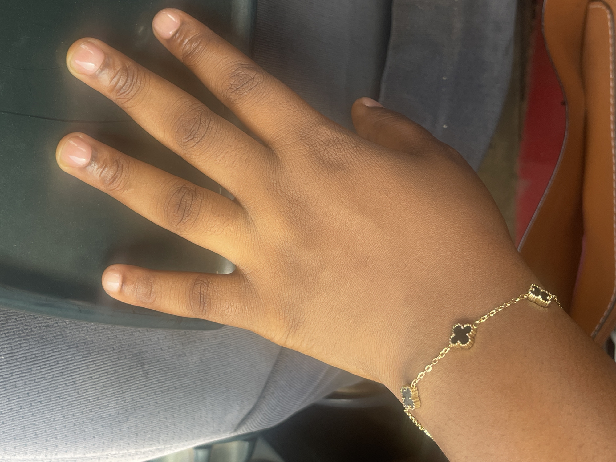 A hand with a gold and black clover bracelet is resting on a gray surface. The background contains a tan purse. There is no evidence in the image to suggest this picture was taken in Nyanya, Nigeria.
