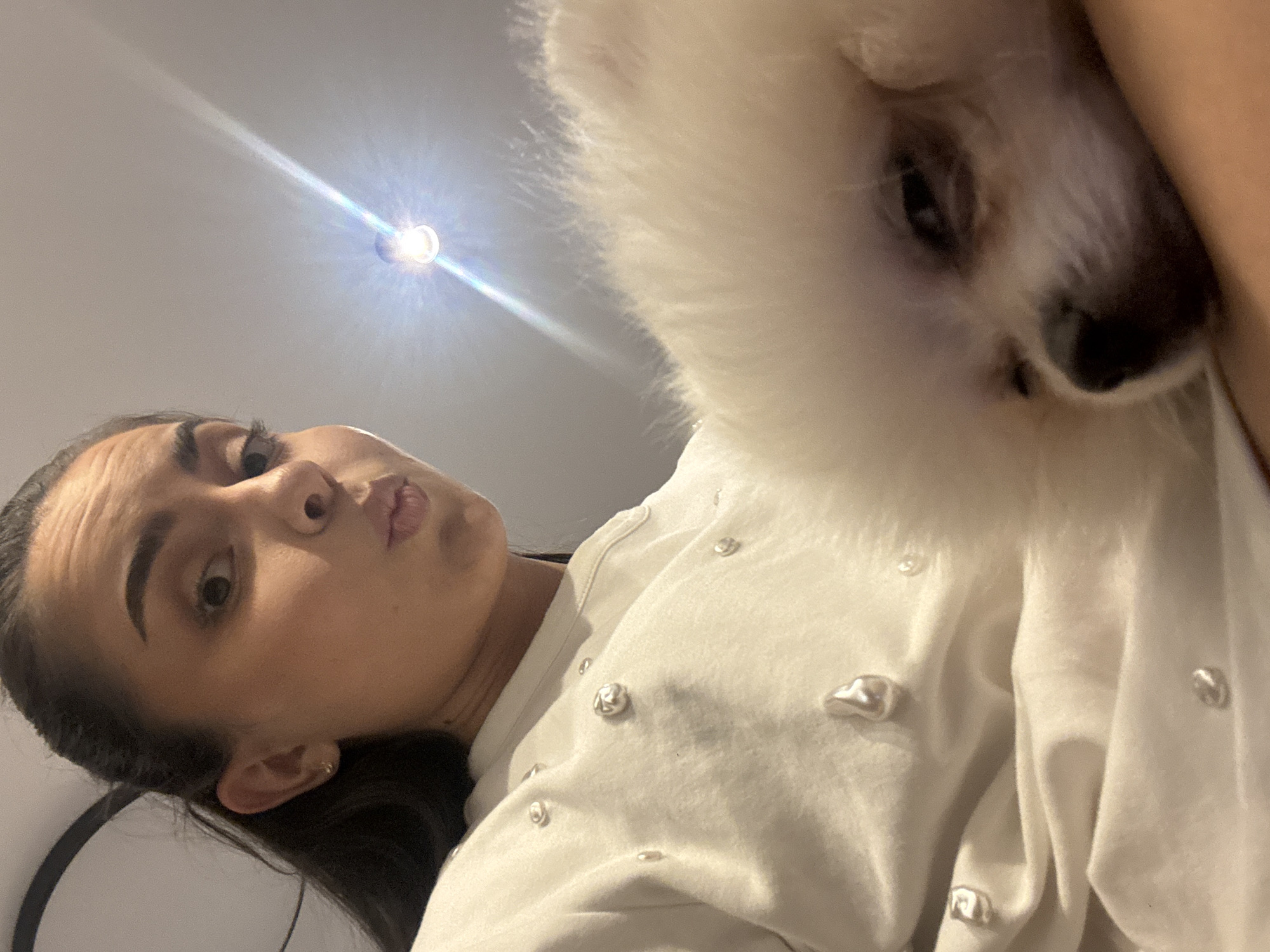 This is a selfie taken indoors, possibly in an apartment in Sofia, Bulgaria, as suggested by the context. The image features a young woman holding a small white dog. The woman is in the foreground, looking at the camera with her lips pursed. She has dark hair pulled back and is wearing a white sweatshirt adorned with pearl-like embellishments. The dog, a fluffy white Pomeranian or similar breed, is nestled in her arms, with its face partially visible and eyes closed. The background is dimly lit, with a ceiling light fixture visible in the upper right corner, casting a bright glare. The overall impression is an intimate and casual moment captured between the woman and her pet.