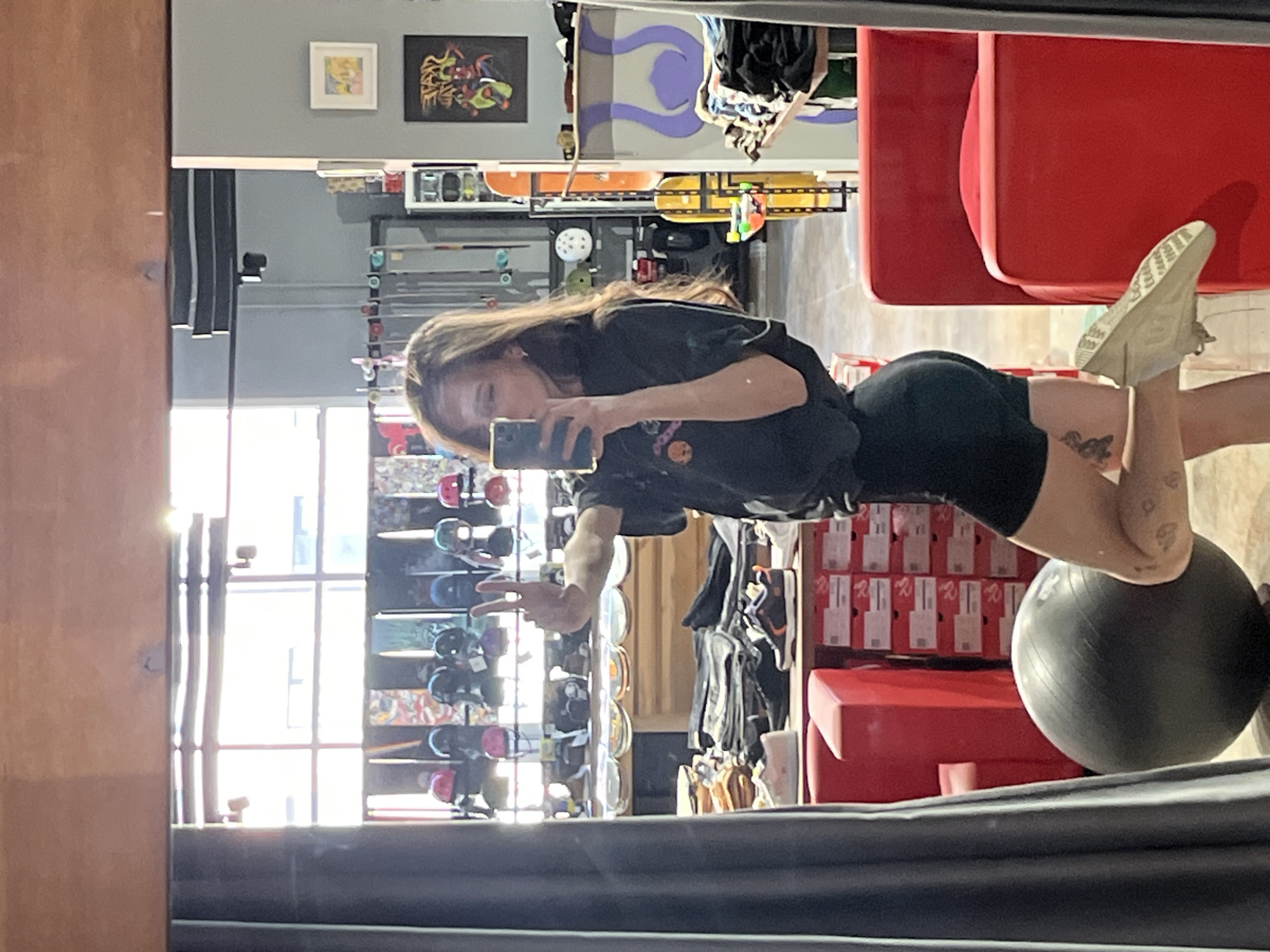 A woman is reflected in a mirror, taking a selfie in what appears to be a retail store. She is wearing a dark t-shirt and black shorts and is balancing with her legs on a gray exercise ball and gesturing with her left hand. Behind her, reflected in the mirror, are rows of skateboards on display, stacks of shoe boxes, and what appears to be gym equipment. The reflection also shows a window with multiple panes. Without further context, identifying the location in Krasnoyarsk, Russia, is not possible.