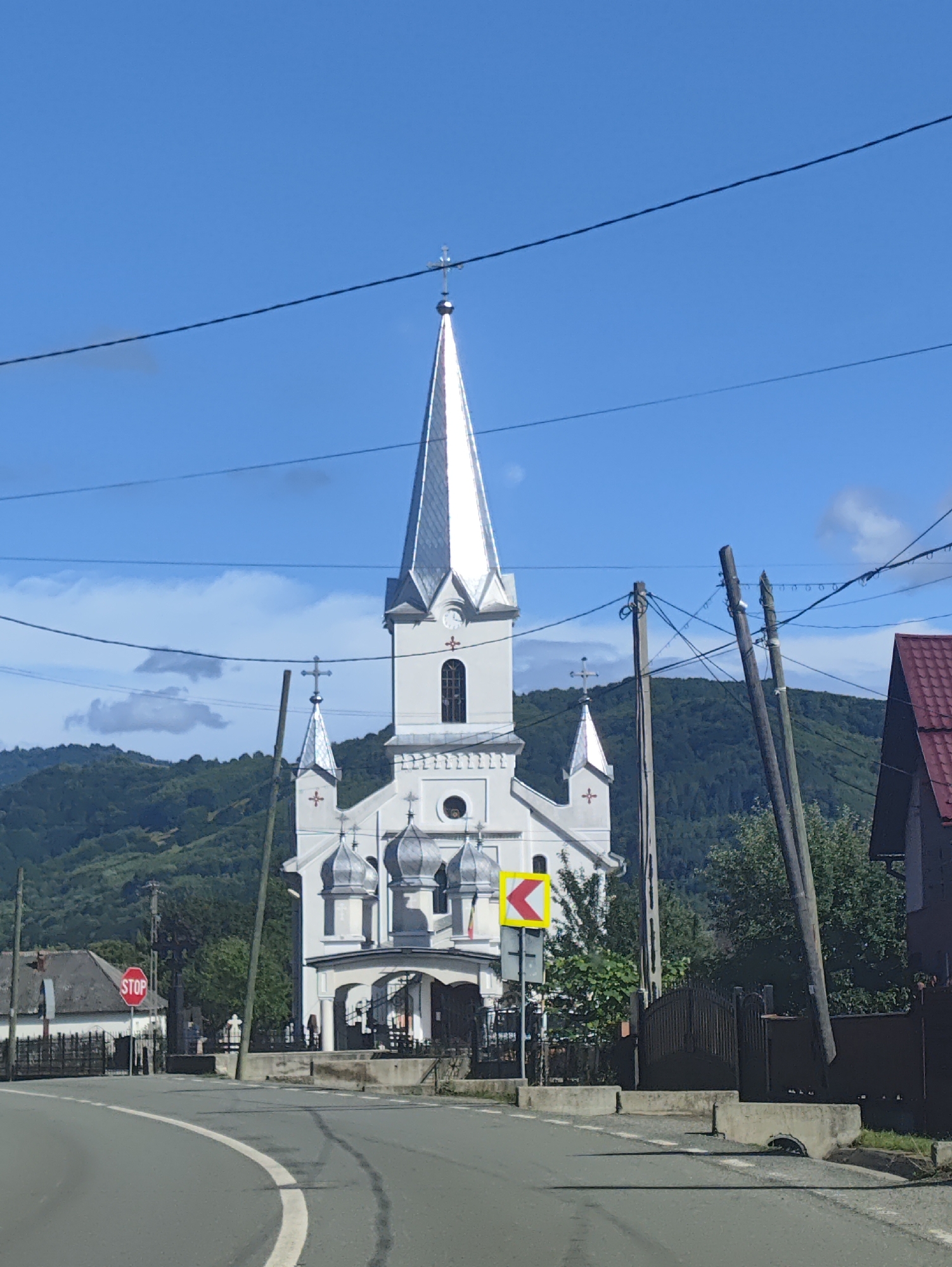 This image depicts a bright, sunny day in a village setting, likely in Leordina, Romania, given the context. The primary subject is a striking white church with a tall, silver spire topped with a cross. The church has several smaller domes and decorative elements, suggesting a traditional architectural style.
In the foreground, a paved road curves to the left, with a white lane marker. A red octagonal "STOP" sign is visible on the left side of the road, near a black metal fence and a small building. To the right of the church, there is a dark wooden fence and a building with a red roof.
Several utility poles with numerous wires crisscross the sky, obscuring parts of the background. Behind the church, rolling green hills covered in dense forest rise under a clear blue sky with scattered white clouds. The lighting and clear sky suggest it is midday.
The scene is peaceful and pastoral, with no people immediately visible. The overall impression is one of a serene rural community. No other legible text is present in the image besides the "STOP" sign.