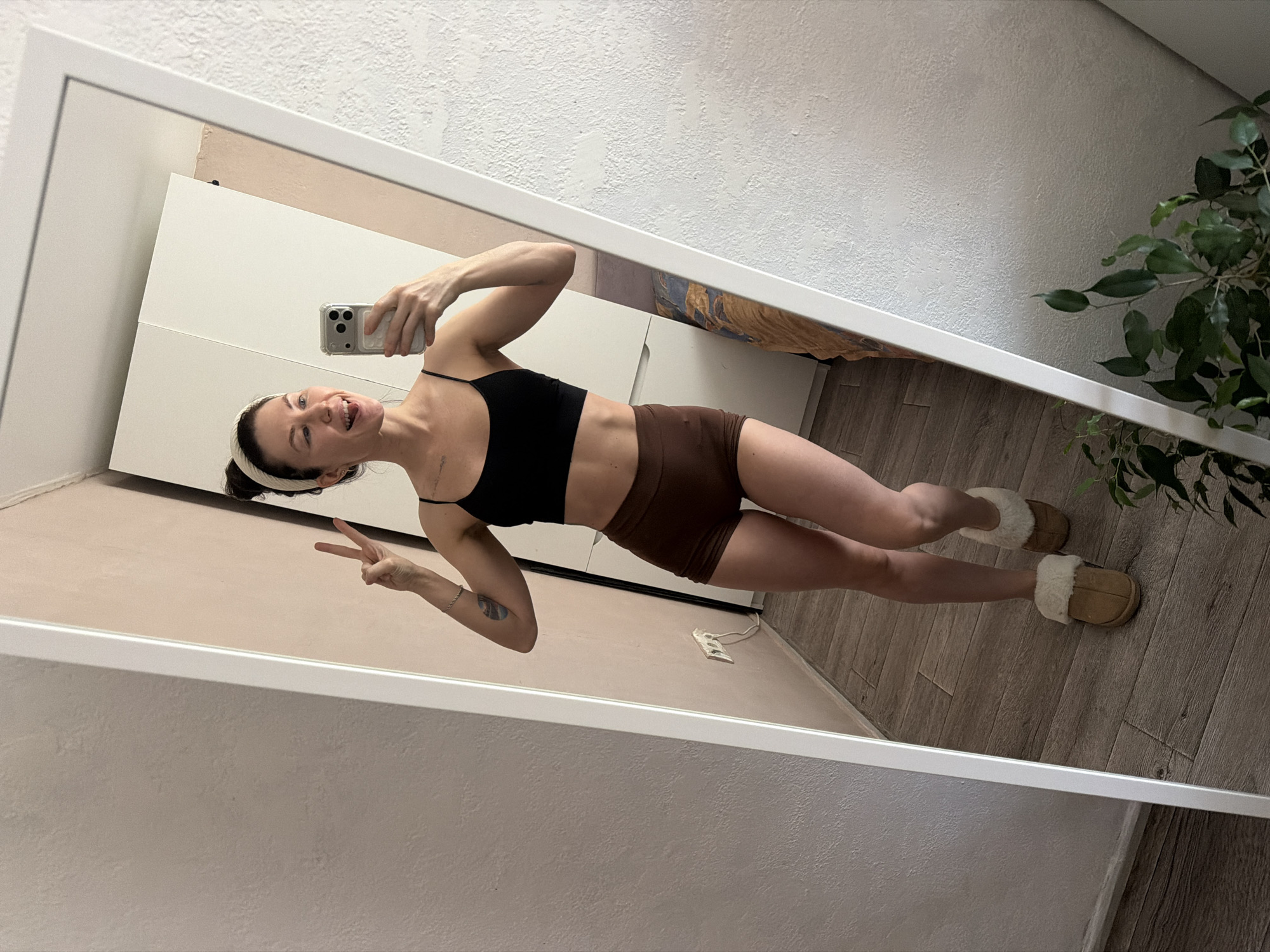 A woman is standing indoors, taking a selfie in a mirror. She is wearing a black sports bra and brown shorts, and slippers. She has a white headband. She is holding her phone in her right hand and making a peace sign with her left hand. In the background is a white cabinet, a wall, a plant and a power outlet. The location is likely a home or apartment. There is no information to confirm that this photo was taken in Perm, Russia.