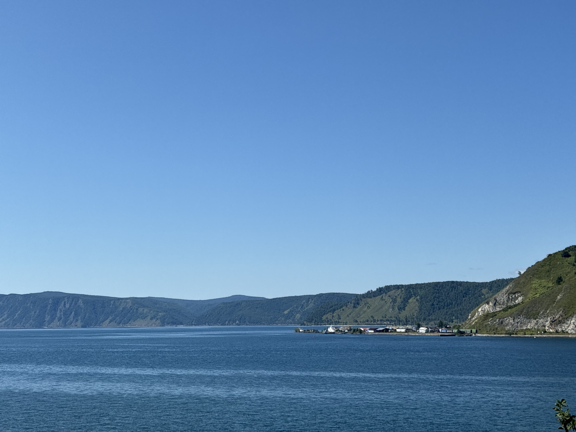 The image depicts a panoramic view of a vast, calm body of blue water, likely Lake Baikal, on a clear, sunny day. The water extends from the foreground towards a distant shoreline. Along the shoreline, a range of rolling, tree-covered hills and mountains forms a natural backdrop, with some areas showing rocky outcrops. In the middle ground, a small settlement is visible, characterized by a collection of buildings with red and white roofs clustered together. Several boats are moored near the shore, suggesting a harbor or a small port. The sky above is a uniform, bright blue, with no clouds present. The overall scene conveys a sense of tranquility and natural beauty, with the settlement appearing to be integrated into the scenic landscape. The location is consistent with the town of Listvyanka, Russia, which is situated on the southwestern shore of Lake Baikal.