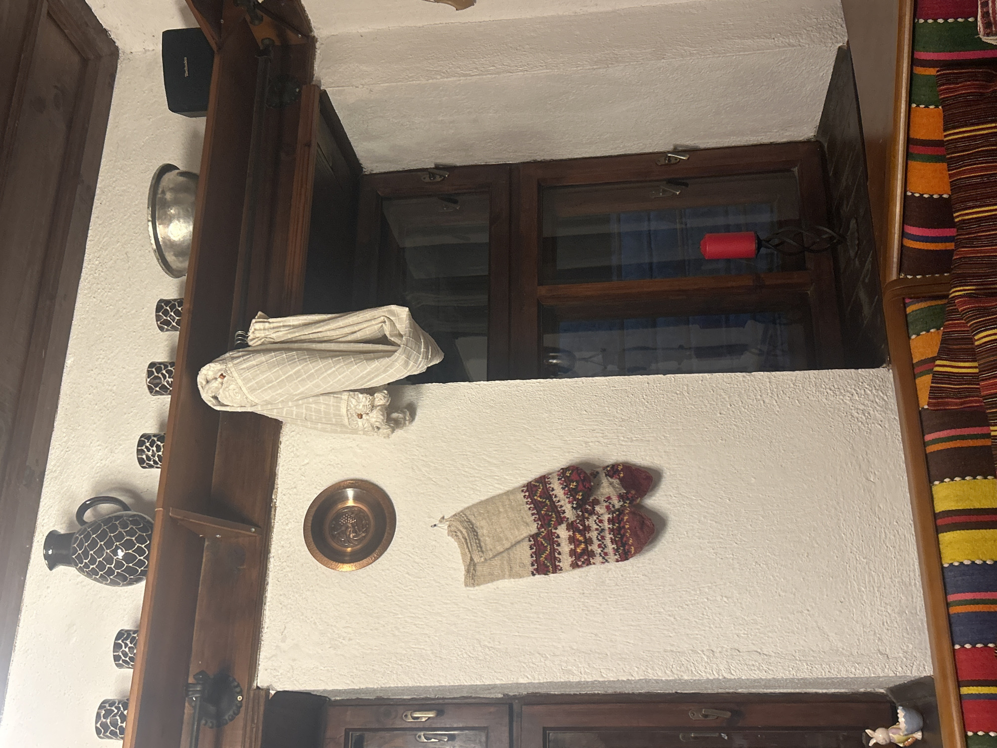 This is an interior shot of a room, potentially a house, in Dolen, Bulgaria. A white wall serves as the primary background. Hanging on the wall are several items: a folded white blanket or cloth, a decorative metal plate, and a pair of knitted mittens with red and beige patterns. The wall also features what appears to be a decorative vase with black and white patterns, mounted on a wooden shelf. The window has a red handle. A colorful striped textile or rug is partially visible on the right edge of the frame. The overall style suggests a traditional or rustic Bulgarian interior.