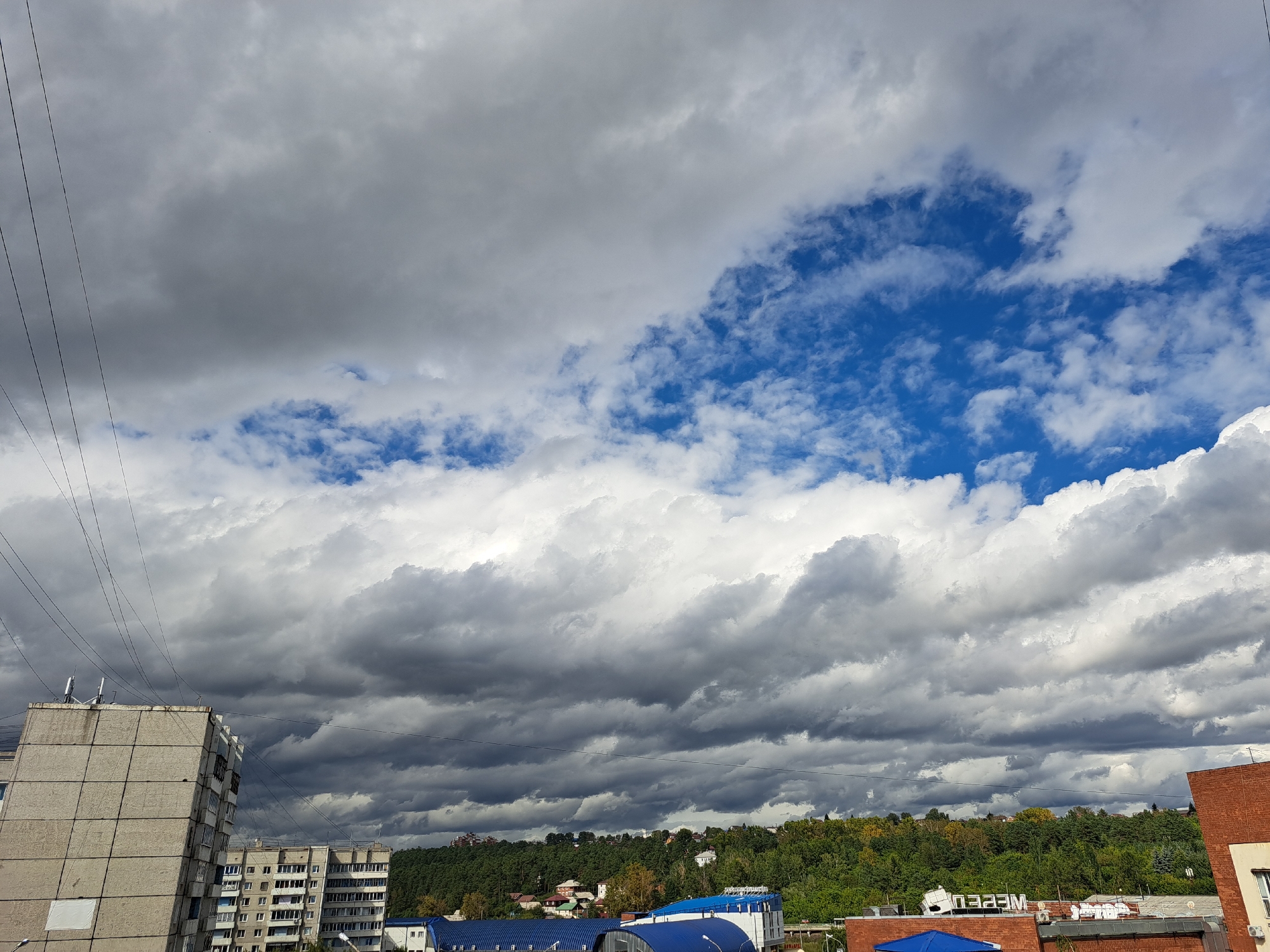 This is a wide-angle, exterior shot looking up at a cloudy sky over a city. The foreground shows the tops of several multi-story buildings, some appearing to be residential with balconies and windows. One building on the left is a light-colored concrete structure with a visible white square on its side. Another building to the right is made of red brick. Overhead, a dense canopy of gray and white clouds fills most of the frame, with patches of bright blue sky visible in the upper right quadrant. Several thin black wires stretch across the upper left portion of the image. In the background, a lush green hillside covered in trees is visible, dotted with houses. The overall impression is of an overcast day in an urban environment, possibly in the city of Irkutsk, Russia, given the architectural style and vegetation. No people or animals are visible in the image.