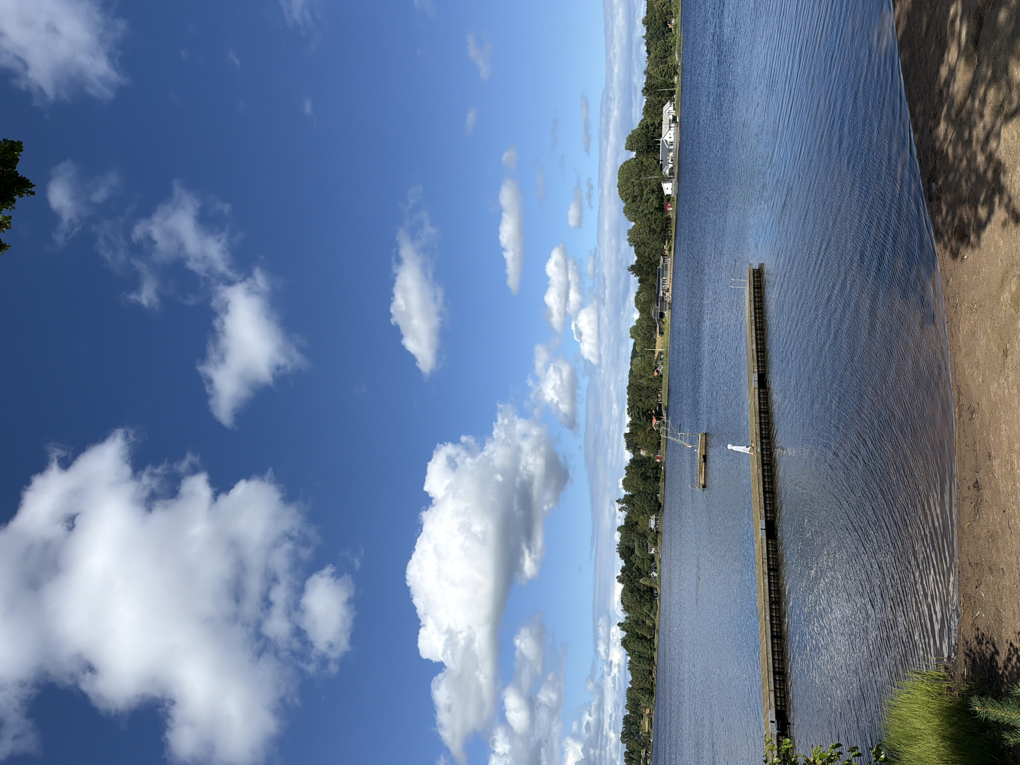 The image depicts a serene lake scene under a partly cloudy blue sky. In the foreground, a sandy beach transitions into the calm, rippling blue water of the lake. A wooden pier extends from the left side of the frame into the water. A lone figure, dressed in white, walks along the pier. In the mid-ground, a small floating platform with a ladder is visible. The opposite shore is lined with dense green trees and scattered houses, suggesting a residential area bordering the lake. The overall atmosphere is peaceful and evokes a summer day. While no specific landmarks are identifiable in the image to pinpoint a precise location within Eksjö, Sweden, the scene is consistent with the natural landscape found around the city's lakes.