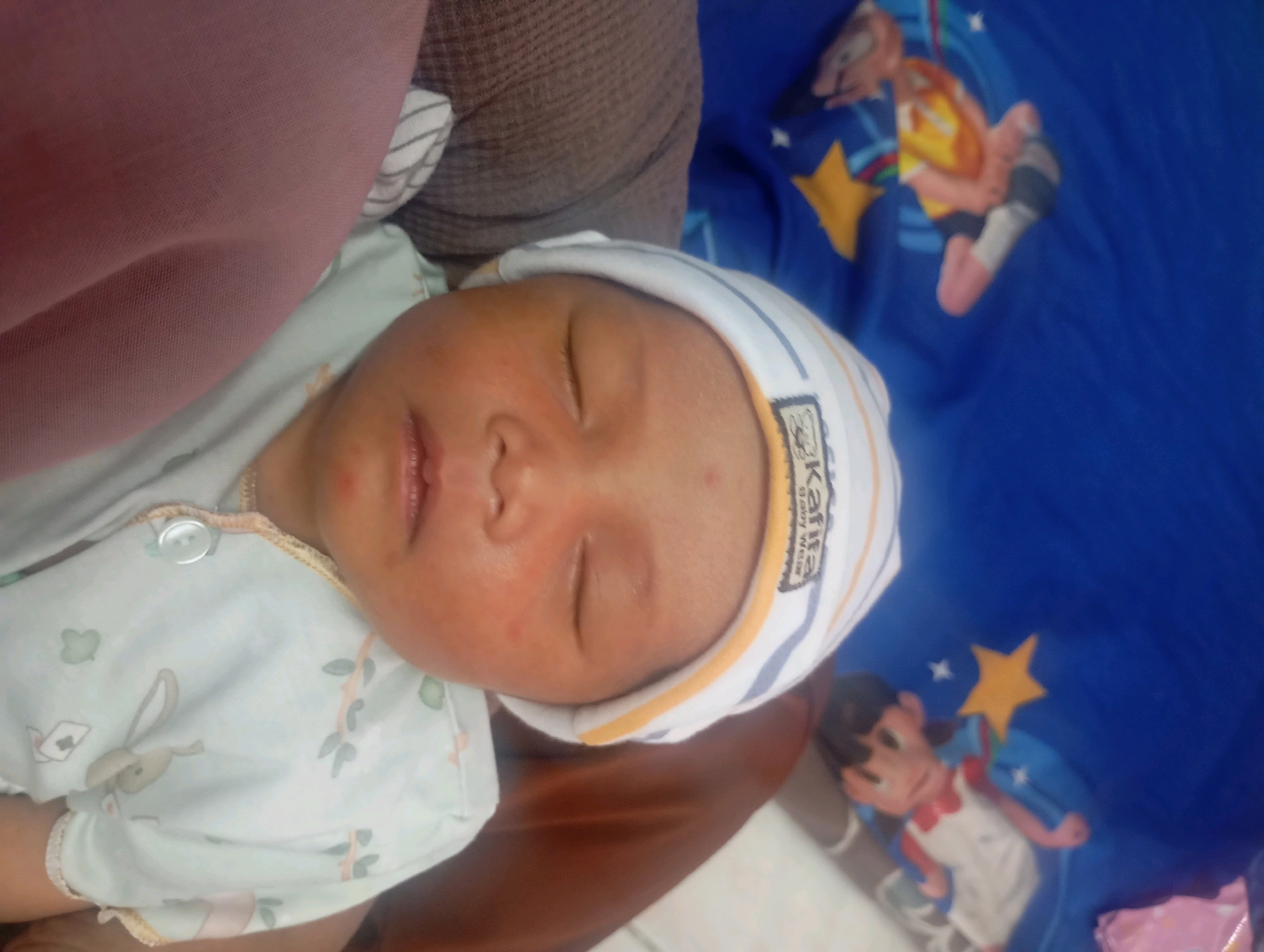 A high-angle, close-up, indoor shot shows a newborn baby sleeping on its back. The baby is wearing a light blue onesie with a white button at the neck and a light blue hat with white and yellow stripes. The hat has a black and white logo on it. The baby's face is turned to the left, and its eyes are closed. The baby has reddish skin and some small red spots on its face.
To the left of the baby is a pink cloth, possibly a blanket or clothing. Behind the baby and to the right is a blue fabric with cartoon characters printed on it, featuring stars and figures that appear to be from a children's show. The overall scene suggests a domestic setting, possibly a home or a hospital room in Babelan, Indonesia.