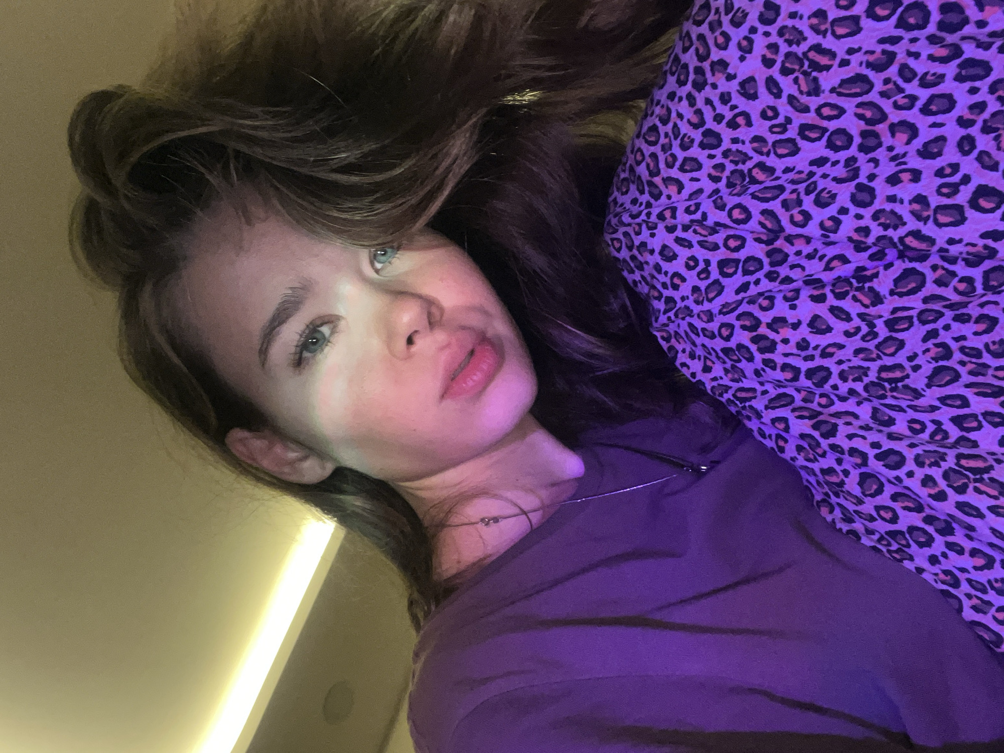 The image features a close-up, diagonal selfie of a young woman with long, wavy brown hair. Her face, including her light-colored eyes (appearing greenish-blue), subtle makeup, and slightly parted lips, is prominent. She wears a plain dark purple t-shirt or top and a delicate silver-colored necklace with small beads. To her right, a significant portion of the frame is filled with a vibrant purple fabric featuring a black and pink leopard print pattern, possibly bedding or another item of clothing.
The activity taking place is a self-portrait or close-up photo.
Notable details include the lighting, which appears to be artificial. A bright, warm-toned linear light source is visible in the upper left background, casting a glow and highlighting aspects of her hair and face. The overall scene has a strong purple and warm yellow/beige color cast due to the lighting. The subject's expression is neutral to slightly contemplative. The setting appears to be indoors, possibly a room, but lacks specific identifiable features such as furniture or architectural details beyond a plain wall and the light fixture. There is no visible text in the image. The indoor setting does not provide visual cues to the "Nam Natural Heritage Area Trang, Vietnam" location context.