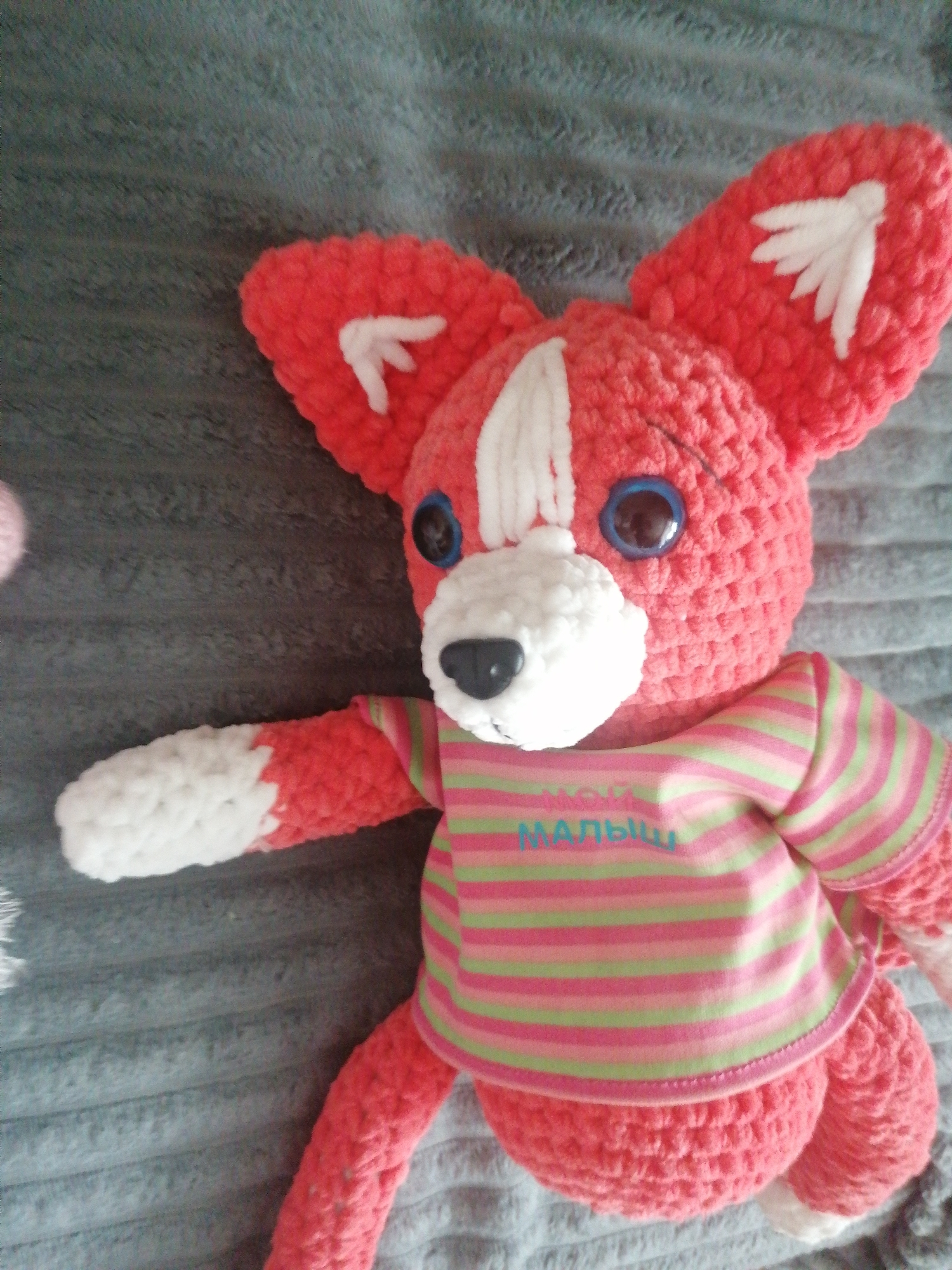 A crocheted stuffed toy resembling a fox or dog is lying on a grey, textured surface. The toy is predominantly bright pink with white accents on its ears, muzzle, and paws. It is wearing a striped shirt with horizontal stripes in pink, light green, and white. The word "МАЛЬЧУ" is embroidered in blue thread across the chest of the shirt. The toy has large, round blue eyes and a black nose. The image does not provide any information about the city of Stavropol, Russia, or any specific location within it.