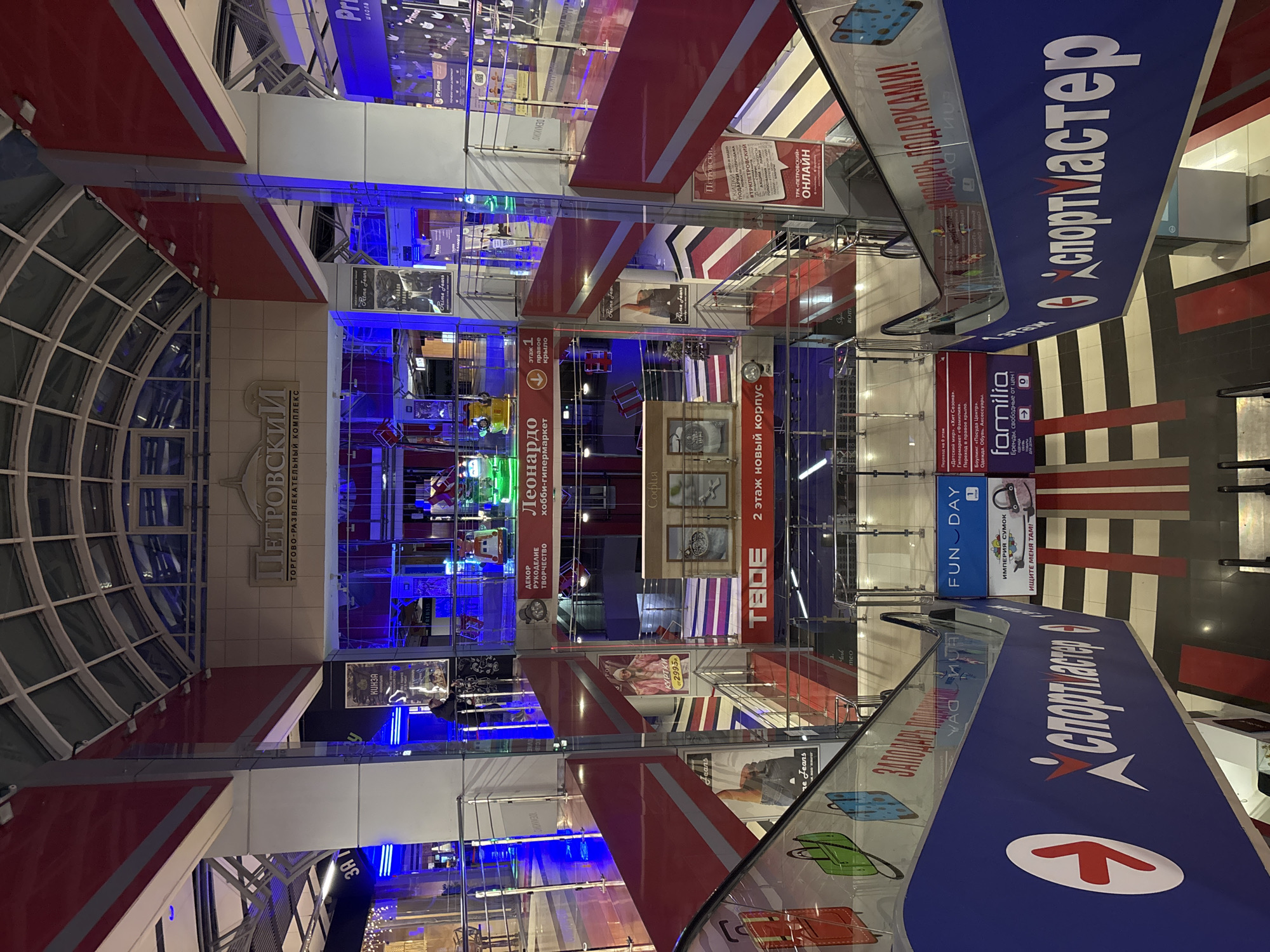 The image depicts the interior of the Petrovsky Trade and Entertainment Complex in Izhevsk, Russia. Visible are multiple levels of the complex, interconnected by escalators and glass railings. Various store signs are visible, including "Спортмастер" (Sportmaster), "Familia," "Леонардо" (Leonardo), and "ТВОЕ" (TVOE). The architecture features a combination of modern design with red and white accents, blue lighting, and a glass ceiling. The image shows a clean and well-maintained environment. There are no people visible in the image.