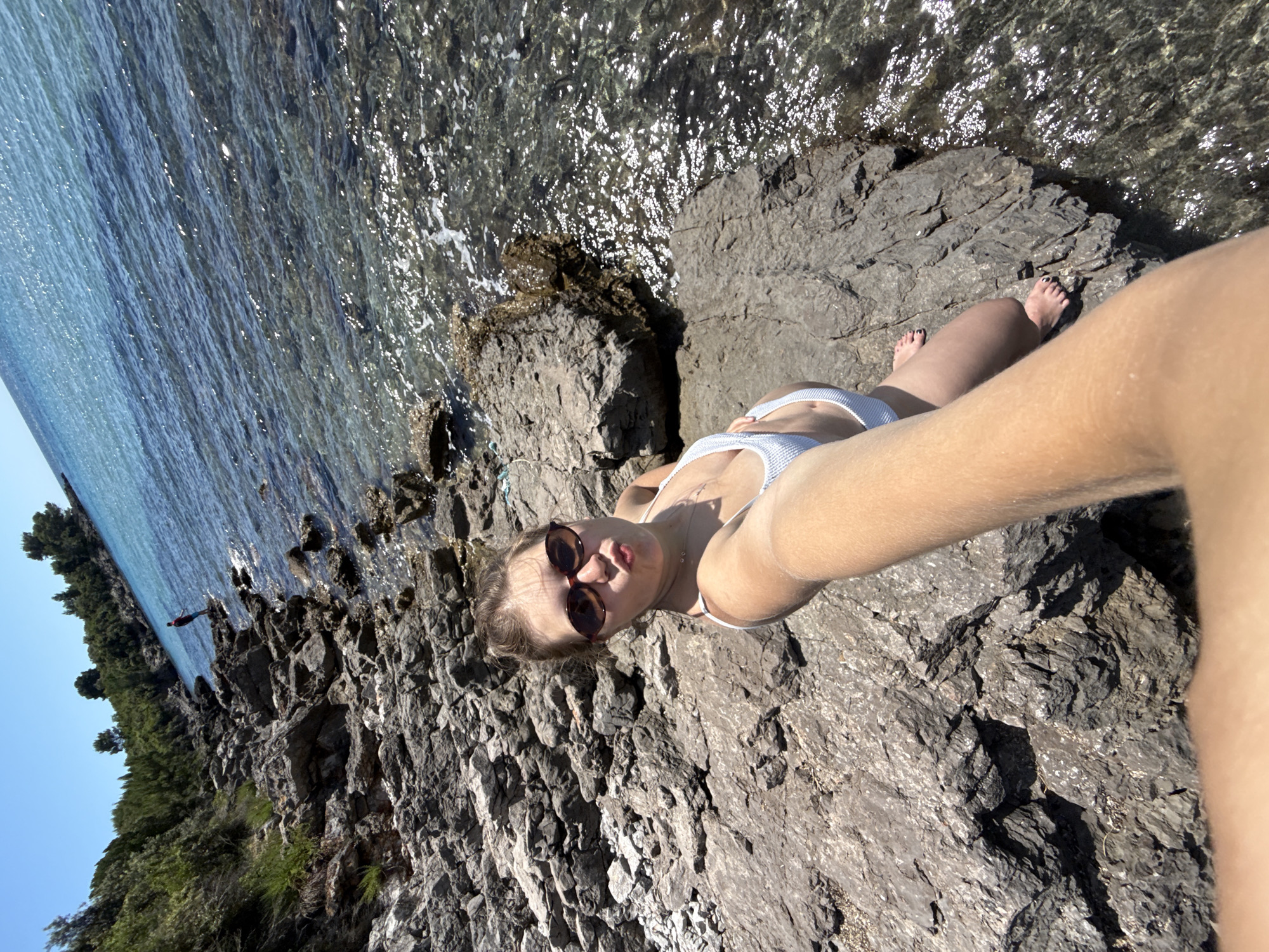 A young woman wearing sunglasses and a light-colored bikini lies on a rocky outcrop beside the Adriatic Sea in Milovići, Montenegro. Her arm is extended, holding the camera in a selfie pose. In the background, a person is diving into the water. The shoreline is visible, lined with trees. The sky is clear.
