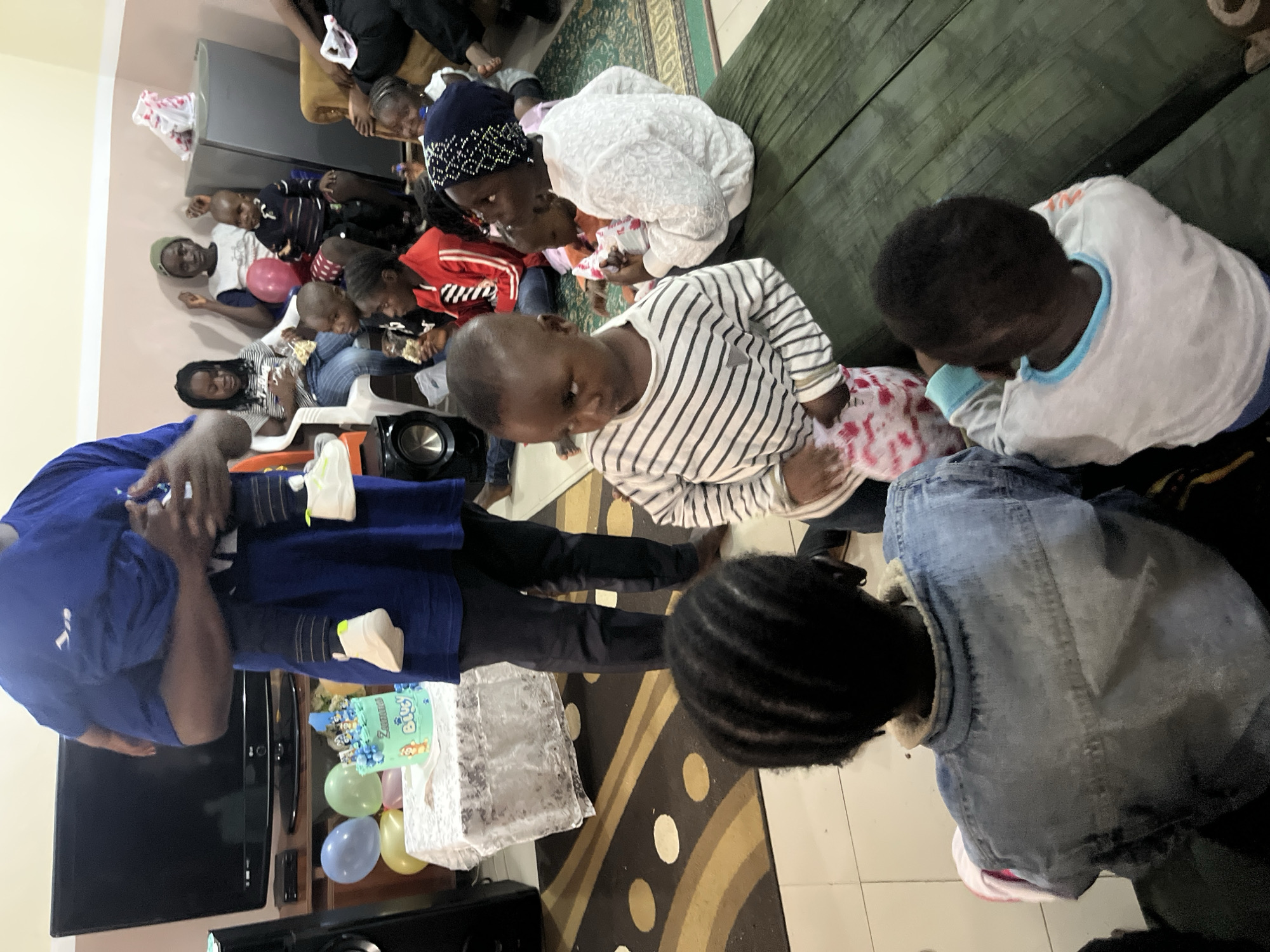 This is a medium shot, eye-level, indoor photograph taken at a birthday party in Bukuru, Nigeria. The scene is a living room, decorated with balloons and a birthday cake. A group of children and adults are gathered, some seated on the floor and others on chairs. In the foreground, a child wearing a blue denim jacket is seen from behind, facing away from the camera. To their right, another child in a striped shirt is looking to the left. A man in a blue shirt is holding a baby, partially obscuring a television set and a table with balloons and a cake. Several other children are visible in the mid-ground and background, some interacting with each other. The overall atmosphere appears to be festive.