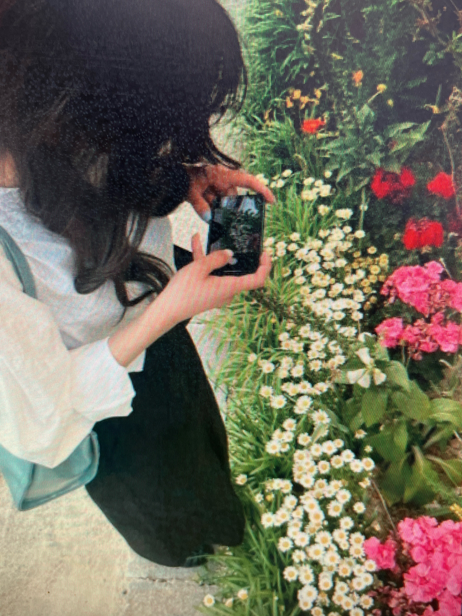 A person with dark hair is taking a photo of a garden with a smartphone. The person is wearing a white blouse and a dark skirt. The garden contains a variety of flowers, including red, pink, and white ones, along with lush green foliage. The ground appears to be a paved surface. The image does not provide any specific information about a location in Sejahtera, Indonesia.