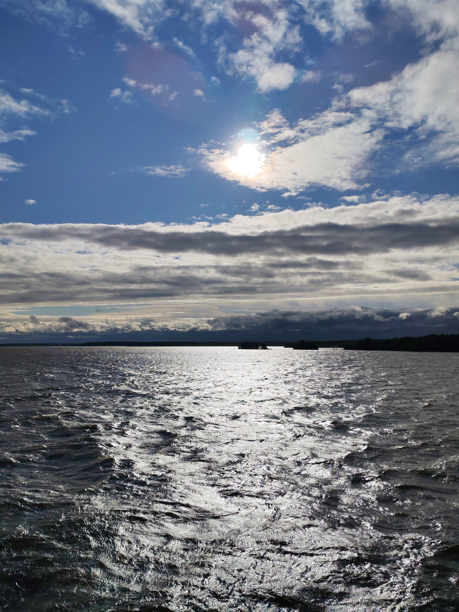 The image depicts a wide body of water, likely a river or lake, under a dramatic sky. The sun is partially obscured by clouds, casting a bright, shimmering reflection on the rippling water. The water appears choppy, suggesting a breeze. In the distance, a dark, tree-lined shoreline is visible, with a few scattered structures or islands. The sky is a mix of deep blue and layers of gray and white clouds. This scene could be located in Sheksna, Russia, a town known for its waterways.