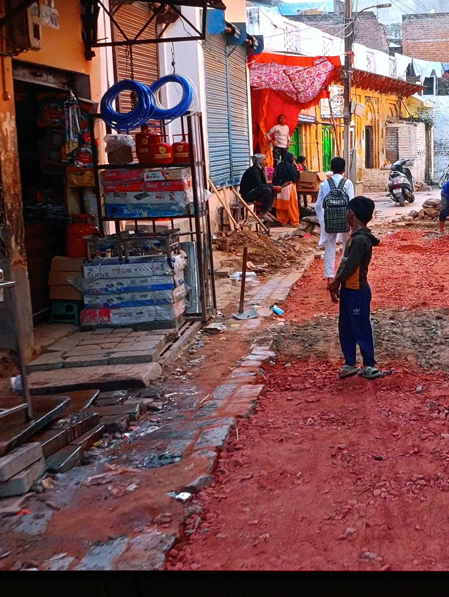 The image depicts a street scene in an urban location, likely in India, characterized by ongoing construction or renovation. The ground is covered with a red, gravel-like material.
In the foreground, a young boy wearing a dark jacket and blue Adidas pants stands with his hands clasped. Behind him, a person with a backpack walks further into the street. Adjacent to the boy, a shovel is stuck in the ground amidst debris and construction materials.
On the left side of the frame is a shop front displaying goods such as blue hosing, gas cylinders, and boxes of "SURYA" labeled products. Several metal trays sit near the shop's entrance.
Further down the street, some individuals are sitting on what appears to be steps or low benches. A scooter is parked nearby. Above them, a red and white patterned cloth is draped, possibly serving as a makeshift awning. Buildings line the street, showing signs of age and wear. A single overhead power line stretches across the sky.