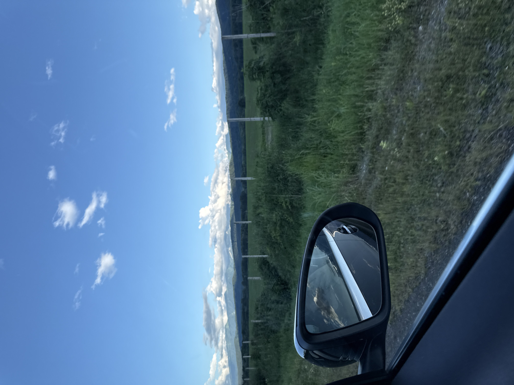 The image, taken from inside a vehicle, shows a view through the side window and the side-view mirror. The side-view mirror reflects the sky, clouds, and the side of the car. Outside the car, a landscape of green fields and vegetation is visible. In the background, a line of trees and mountains can be seen under a bright blue sky with scattered white clouds. A fence line with posts runs horizontally across the middle ground. The overall scene depicts a rural or semi-rural environment. There are no people or specific landmarks to identify the location within the city of Alagir, Russia.