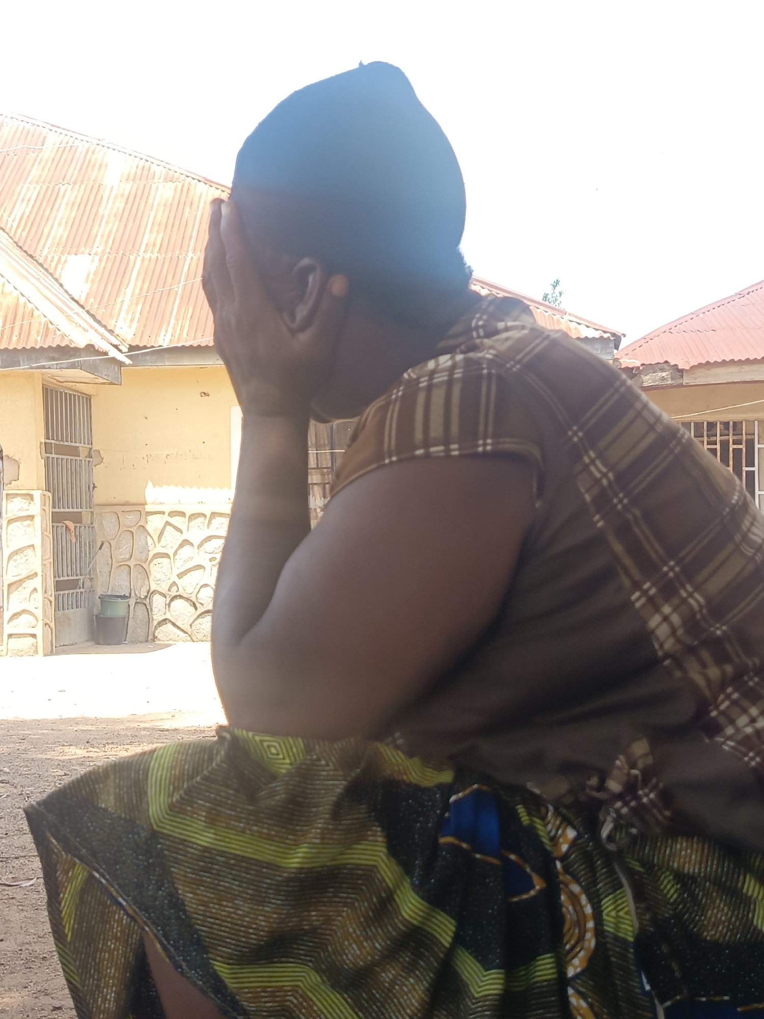 An adult woman in Akwanga, Nigeria is sitting outdoors in front of buildings with corrugated metal roofs and barred windows. She is wearing a dark head covering, a plaid shirt, and a patterned skirt. Her face is obscured by her hands.