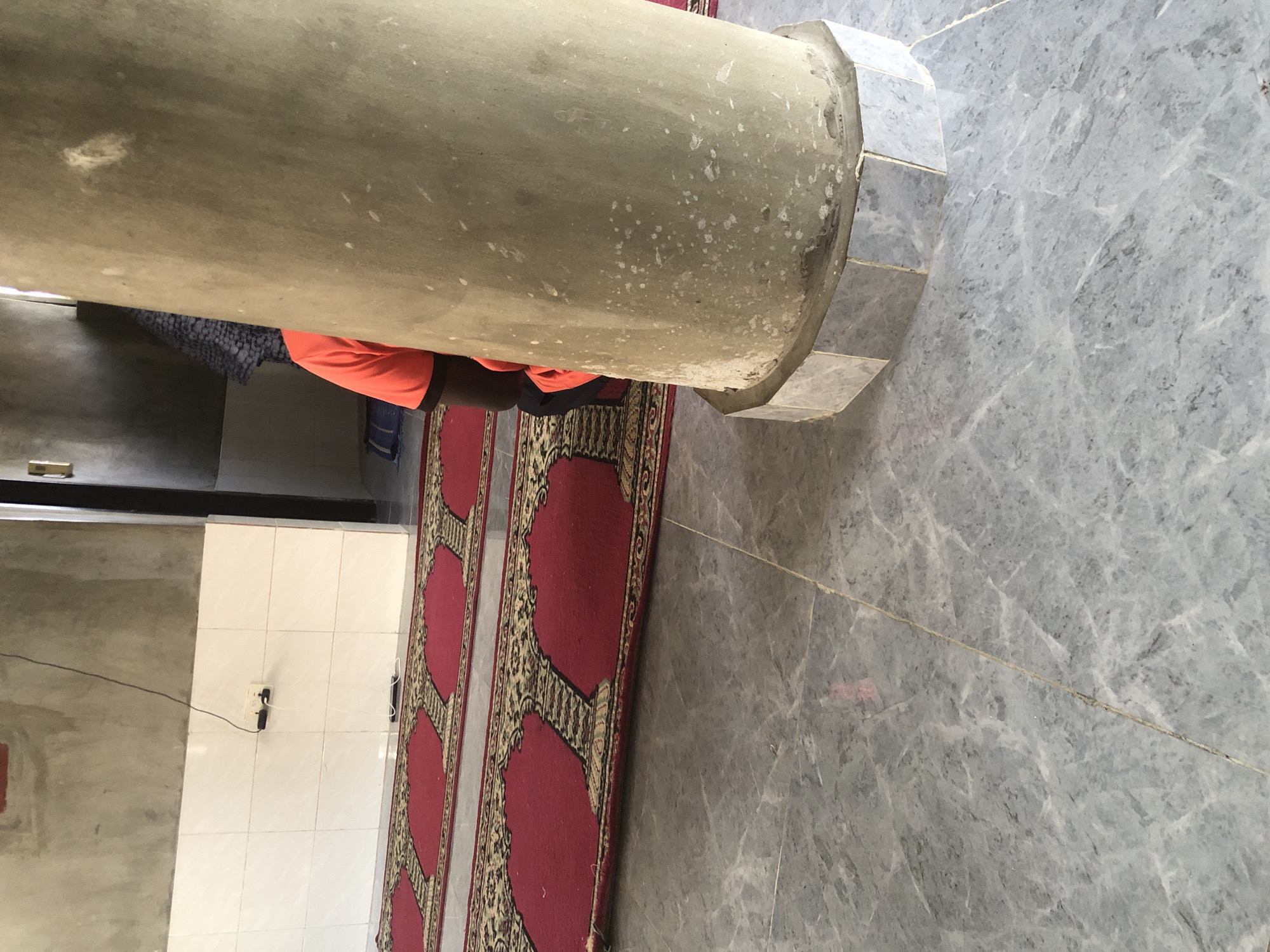 An interior shot captures a person kneeling on a prayer rug. The person, wearing an orange t-shirt, is positioned behind a thick, textured concrete pillar that dominates the foreground and right side of the frame. The pillar has a rough, weathered appearance with visible white speckling. The prayer rug, which is red with a black and gold intricate pattern, is laid out on a grey marble-tiled floor. Two rows of these prayer rugs are visible, extending towards the background. The wall behind the rugs features white tiles on its lower half and a rough concrete finish on the upper half. Electrical outlets are visible on the tiled wall. The background shows a doorway and another pillar, suggesting a larger indoor space, possibly a mosque in Ado, Nigeria.