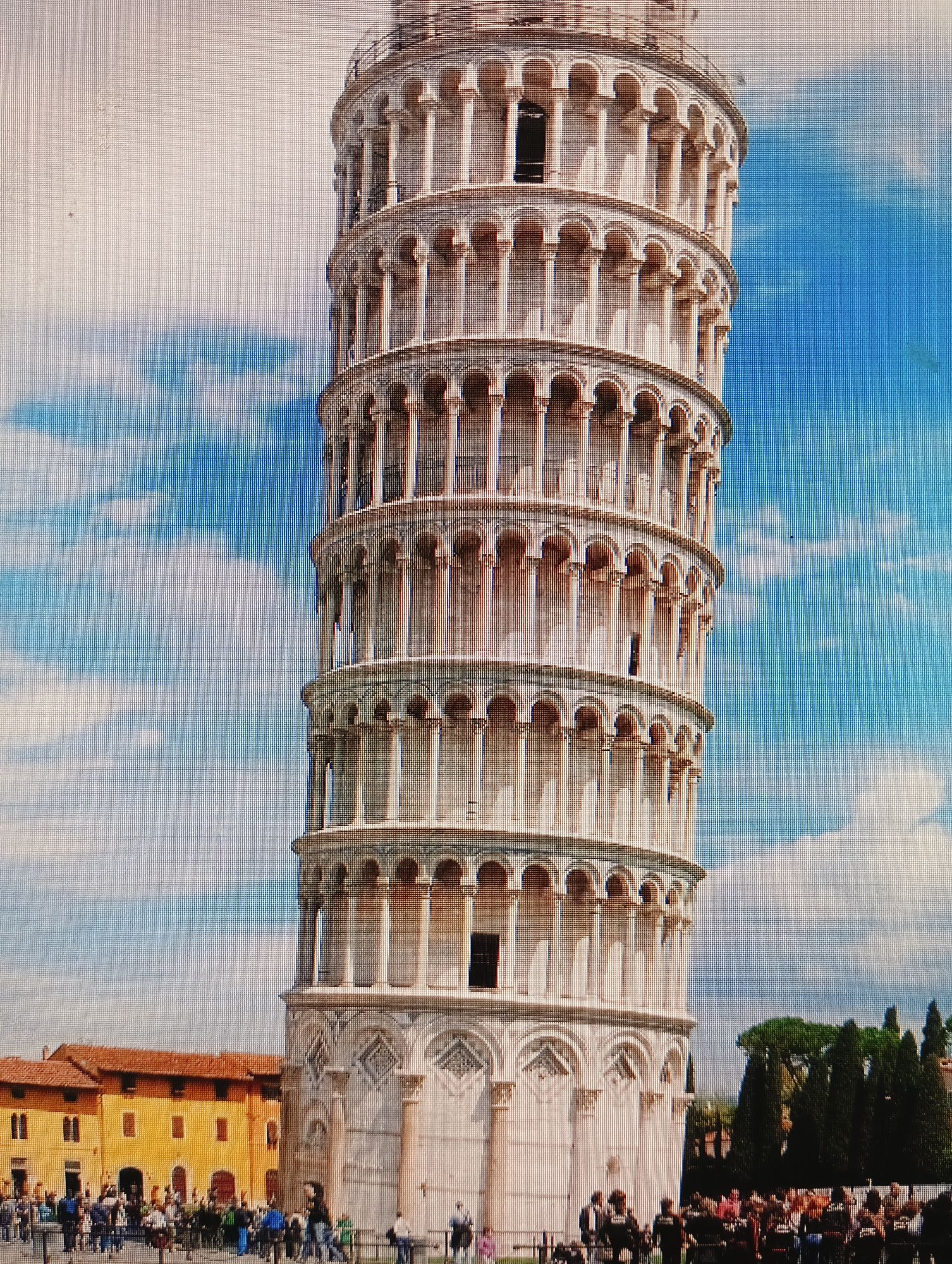 The image shows the Leaning Tower of Pisa. The tower is made of white marble and has a cylindrical shape with arches and columns. The tower is tilted to the right. In the background, there are buildings with orange roofs and green trees. There are many people standing in front of the tower. The sky is blue with white clouds. There is no indication of the city of Sluck, Belarus in this image.