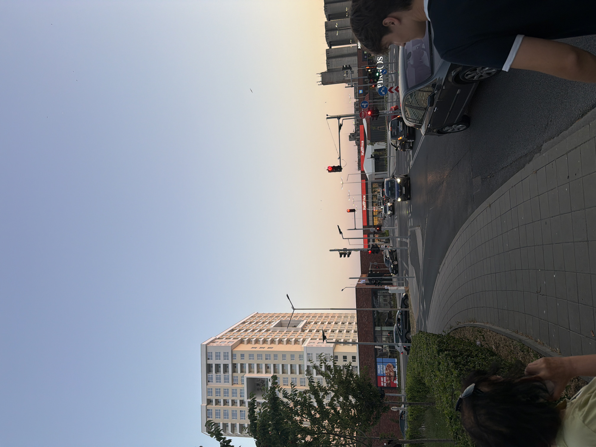 The image captures a street scene in Burgas, Bulgaria, during what appears to be sunset or early evening. The sky is a soft gradient of pale blue and orange, suggesting the end of the day. In the foreground, the tops of people's heads are visible, with one person in a light green shirt wearing sunglasses and another person in a dark t-shirt with a white trim. The perspective is from a low angle, looking up towards the urban landscape.
Dominating the right side of the image is a multi-lane road with several cars. Traffic lights are visible, with some illuminated red, indicating a stop. A dark-colored sedan is prominent, and other vehicles are visible further down the road. A paved walkway with a distinctive curved pattern runs alongside the road, bordered by a low hedge.
To the left, a tall, modern apartment building with a light-colored facade and numerous balconies is the most striking architectural feature. The building has a unique open-air courtyard design in its upper section. Adjacent to the apartment building, and partially visible behind the trees, is a lower, multi-story building with a brick facade, likely commercial or retail, featuring a sign that reads "HalfPrice" twice. Another sign with text and an image of food is also visible, partially obscured by greenery. The presence of traffic lights, cars, and the apartment building suggest an urban residential and commercial area. The overall mood seems calm, with the soft lighting contributing to a peaceful evening atmosphere.
