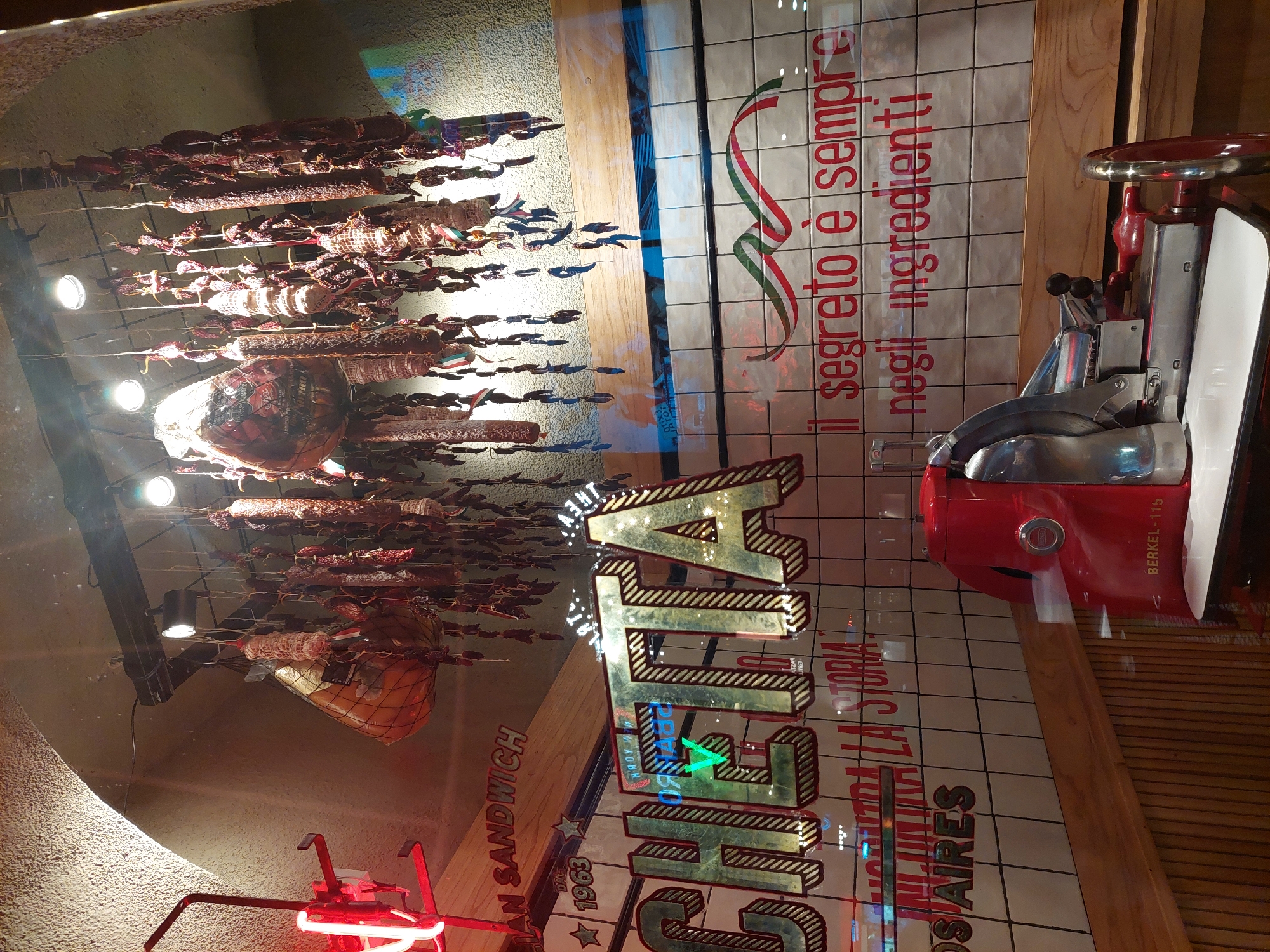 An angled shot captures the interior of what appears to be a traditional Italian deli or sandwich shop, seen through a reflective window. The primary subjects are an extensive display of cured meats and a vintage red deli slicer.
Numerous sausages, salamis, and larger cuts of meat, possibly hams or prosciuttos, hang from metal racks across the ceiling and upper walls. These various types of cured meats are illuminated by several bright spotlights, creating a warm, inviting glow.
To the right, a striking red Berkel-115 manual deli slicer stands on a counter, showcasing a classic design. Behind the slicer, a tiled wall features a decorative ribbon in the green, white, and red colors of the Italian flag.
Visible text on display includes the Italian phrase "IL SEGRETO È SEMPRE NEGLI INGREDIENTI" ("The secret is always in the ingredients"). A large, illuminated sign spells out "CHETTA," which may be part of the establishment's name. Other text references "DAN SANDWICH," "DA 1963" (meaning "Since 1963"), and "LA STORIA" ("The story"), indicating a long-standing business with a focus on sandwiches. The mention of "BUENOS AIRES" explicitly places the location in Argentina, suggesting an Italian-Argentine heritage.
Reflections on the glass surface obscure some details but add to the atmospheric quality, hinting at the outside environment. The scene evokes a sense of artisanal tradition and culinary quality, with no people visible in the frame. The lighting suggests an evening setting or an interior designed for dramatic presentation.