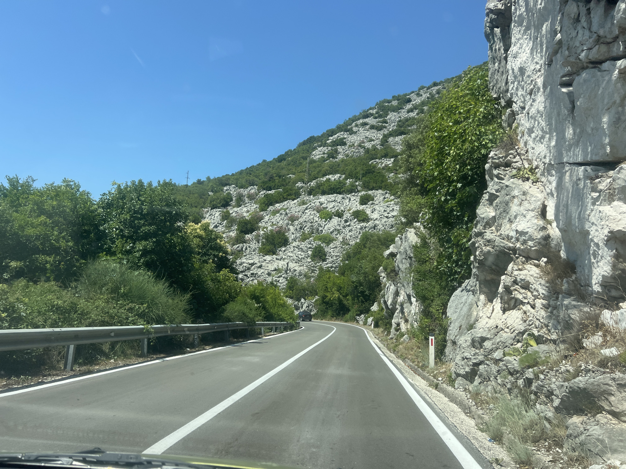 The image captures a winding road in a mountainous, rocky terrain, likely in the vicinity of Štrp, Montenegro. The road is asphalt with white lane markings, bordered by a metal guardrail on the left and a steep, rocky cliff face on the right. Lush green trees and vegetation are scattered across the slopes, interspersed with large grey rocks. The sky is clear and blue. A dark-colored car is visible in the distance on the road. The perspective suggests the photo was taken from a moving vehicle.