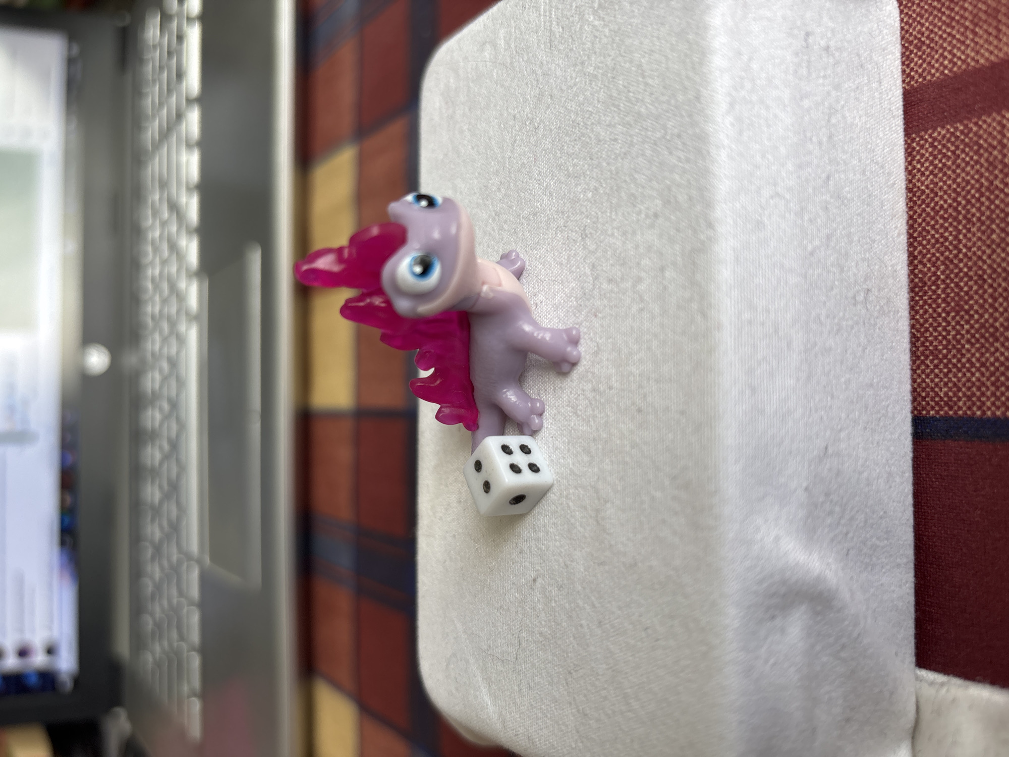 A macro shot focuses on a small, purple toy creature with large blue eyes and pink wing-like appendages, clinging to a white, textured surface. To its right, a white six-sided die rests against the surface, showing four black dots on its visible face. The toy creature appears to be looking towards the viewer with an amused expression. The background is out of focus, showing a dark-colored laptop or electronic device on the left and a reddish-brown and dark blue plaid pattern on the right, suggesting an indoor setting. The lighting is bright, highlighting the textures of the toy and the white surface. No text is visible in the image. The image does not provide specific cues to pinpoint the exact time of day or weather conditions. The setting appears to be a desk or personal workspace.