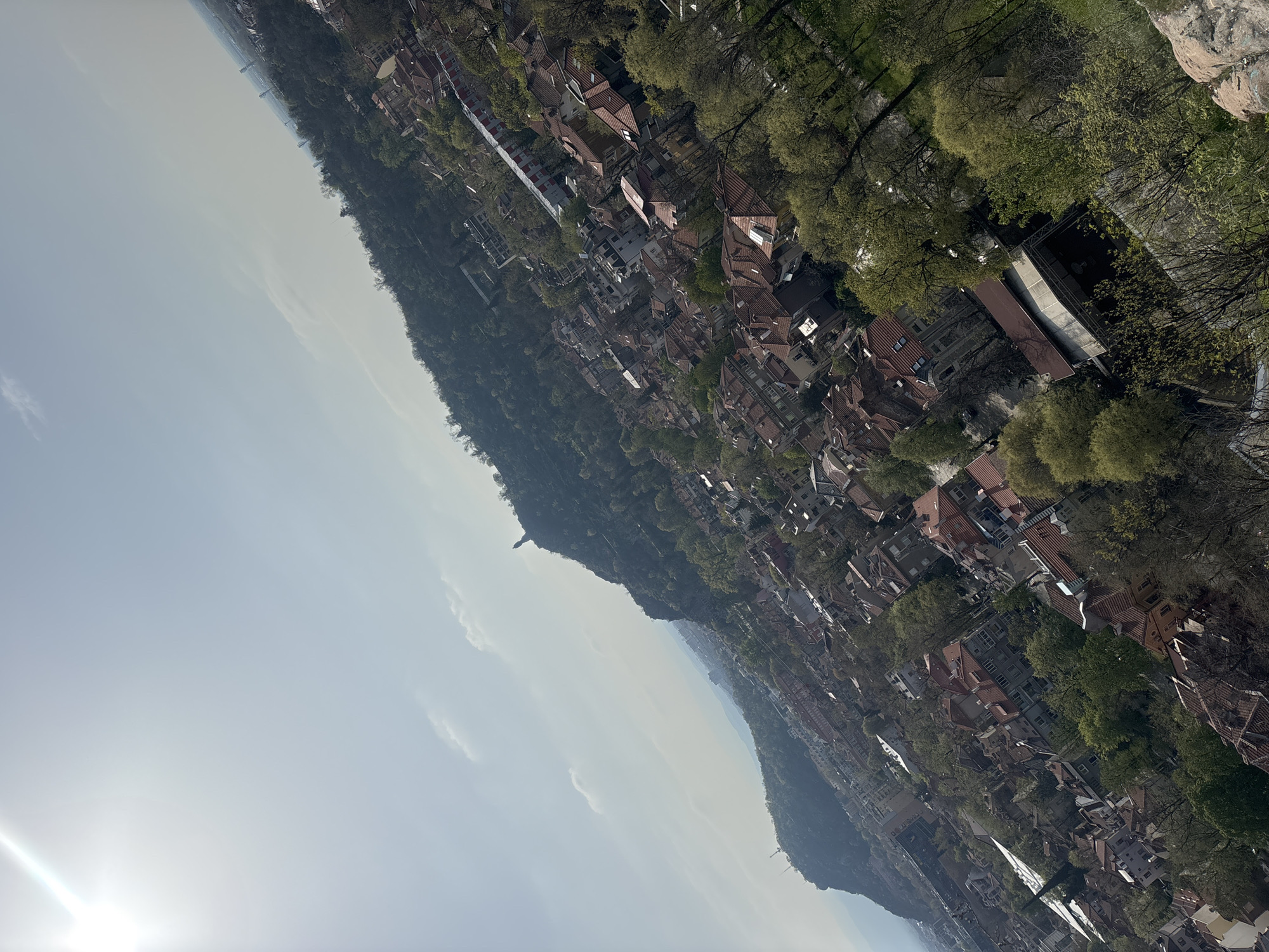 The image is an aerial view of a city, likely taken from an elevated position like a hill or mountain. The scene is dominated by a dense cluster of buildings with red-tiled roofs, interspersed with trees. The architectural style suggests a European setting.
A prominent hill or mountain range runs along the central area of the image, partially covered with trees. The foreground shows a clear sky with some light cloud formations. The sun is visible at the bottom left, casting a hazy light across the scene.
Specific details identifying the location as Plovdiv, Bulgaria are not discernible without additional visual or contextual clues.