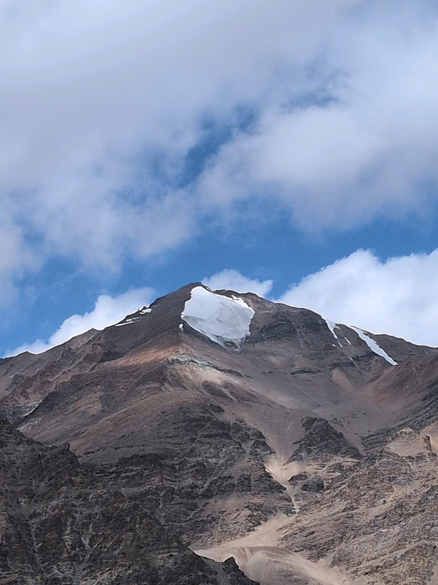 An upward-facing, close-up shot captures a snow-capped mountain peak under a blue sky with scattered white clouds. The mountain is rugged and brown, with a prominent white snowfield on its summit. The texture of the mountain is rough, showing layers of rock and scree. The sky is a vibrant blue, contrasting with the white of the snow and clouds. The lighting suggests it is daytime, with the sun illuminating the mountain from an unseen angle. The image appears to be taken in a remote, mountainous region, with no signs of civilization.