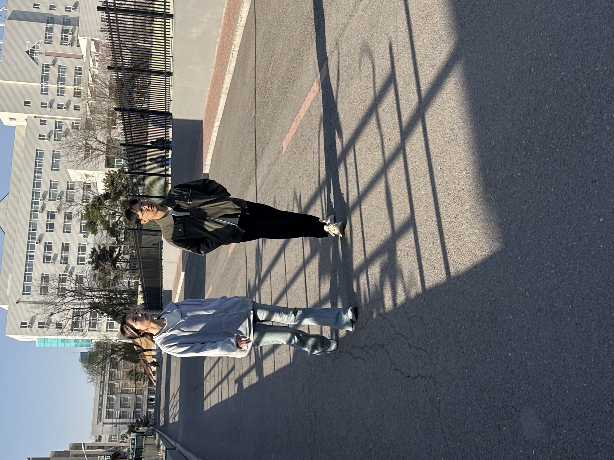 Two people are standing on a paved road. A person wearing a blue hoodie and jeans is standing to the left of a person wearing a green jacket and black pants. Behind them are tall buildings. The shadow of a fence is cast on the road. The location is Zhongtun, China.