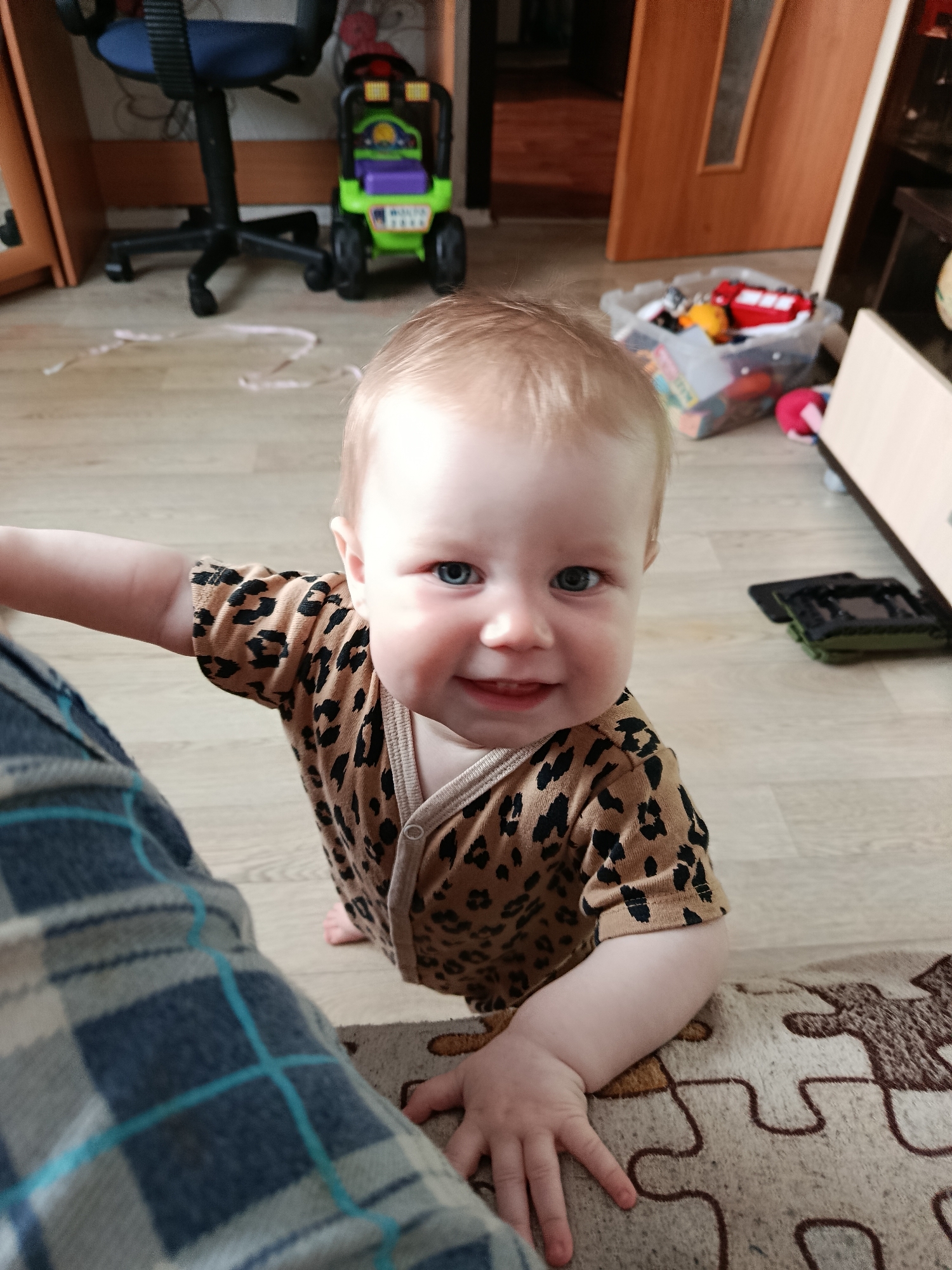 A heartwarming close-up shot captures a baby looking directly at the camera, smiling broadly. The baby has light hair and bright blue eyes, and is wearing a leopard print onesie. They appear to be leaning or crawling towards the viewer, with their right hand resting on a patterned rug or blanket, and their left arm extended towards a person whose leg, covered in a blue and grey plaid fabric, is visible in the lower left foreground.
The scene is an indoor setting, likely a home, with a light wooden floor. In the background, various household items and toys are visible. To the left, there's a black office chair with a blue seat and backrest, and behind it, a small green toy car or truck with purple accents. To the right, a clear plastic bin filled with colorful toys, including a red bus, can be seen. Further back, a wooden door with a vertical frosted glass panel is visible, suggesting a room beyond. The overall lighting indicates it is daytime. The baby's joyful expression is the prominent emotional element of the image. No visible text is discernible.