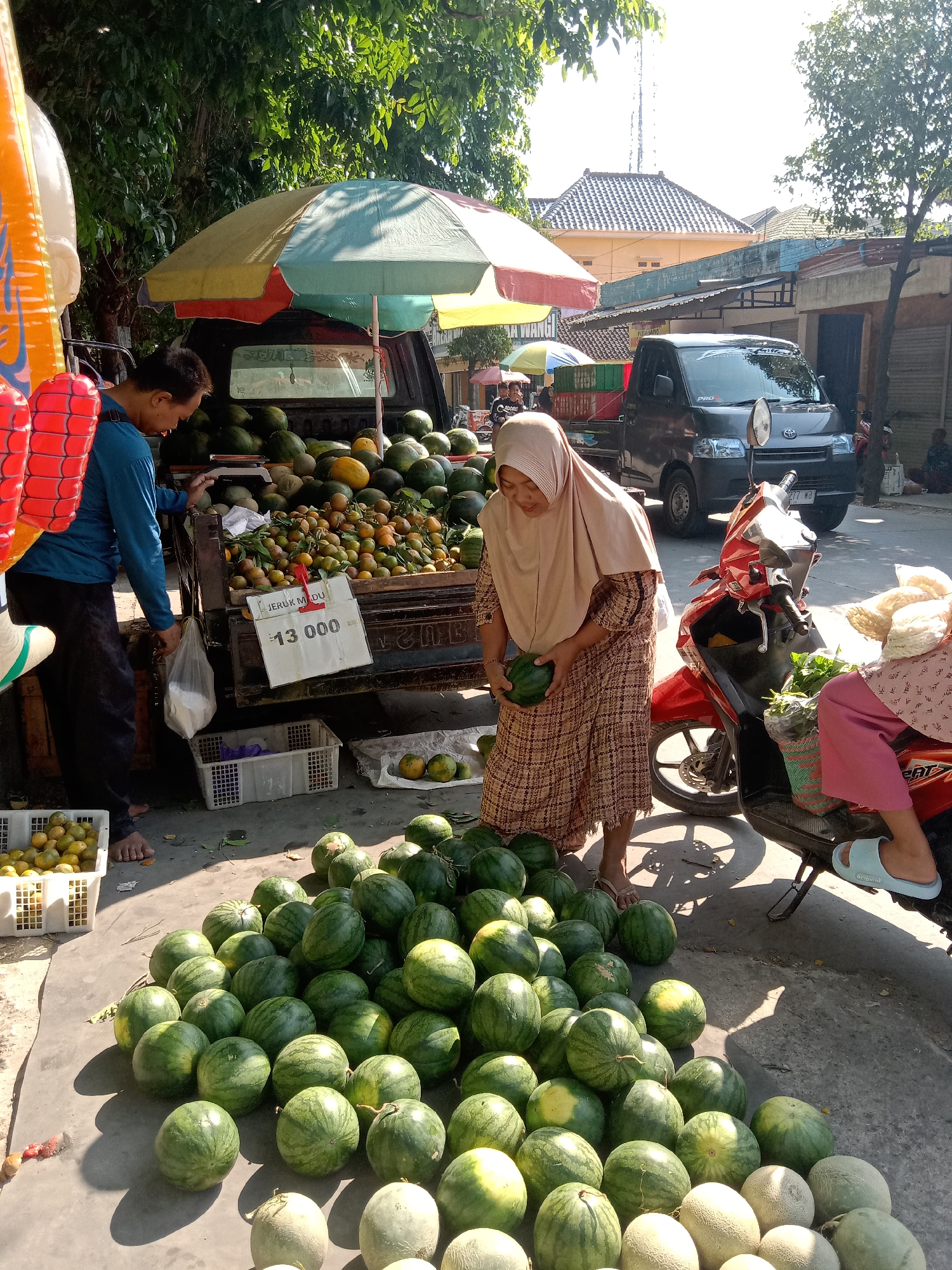 In Sugihwaras, Indonesia, a bustling street scene unfolds with a fruit stall as the central focus. Sunlight streams down, casting sharp shadows on the paved ground. A man, wearing a blue long-sleeved shirt and dark pants, stands next to a pickup truck filled with watermelons and oranges. He holds a plastic bag in his left hand. To his right, a woman in a light brown hijab and a patterned dress examines a small watermelon she holds in her hands.
The pickup truck, a dark gray model with a "PRO" decal on its rear door, serves as the primary display area for the fruits. Large piles of watermelons are stacked in the truck bed and also spill onto the ground in front of it. A vibrant, multi-colored umbrella provides shade over the truck and the fruit display. To the left of the truck, a white plastic crate overflows with oranges, and a red and yellow inflatable decoration hangs nearby.
Adjacent to the fruit stall, a red scooter is parked. A woman, partially visible, sits on the scooter wearing a pink outfit and light blue sandals. She appears to be interacting with a basket of green vegetables. Various white plastic crates are scattered around the stall, some empty and some containing fruit. In the background, buildings with tiled roofs suggest a typical Indonesian urban environment. The overall atmosphere is one of commerce and daily life in a sun-drenched Indonesian town.