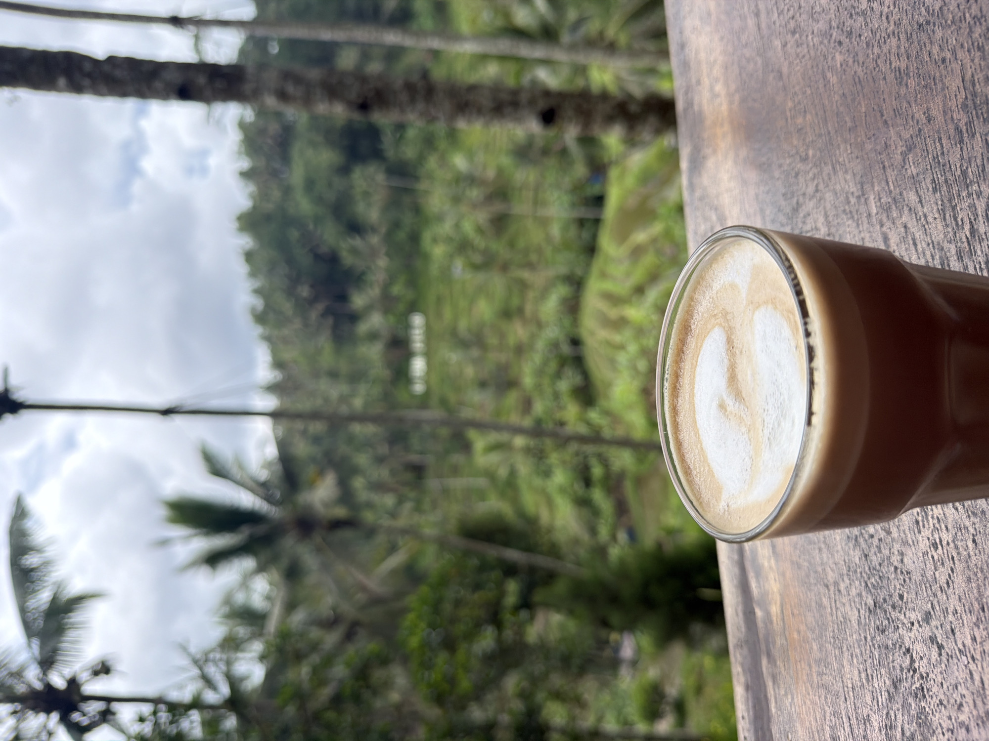 An outdoor shot shows a glass of coffee on a wooden surface with a blurred background of rice terraces and foliage, likely in Tegal Suci, Indonesia. The coffee has latte art, a heart-like shape. The wooden surface has visible grain. The background consists of a verdant landscape featuring rice terraces, trees, and foliage. The sky appears overcast.