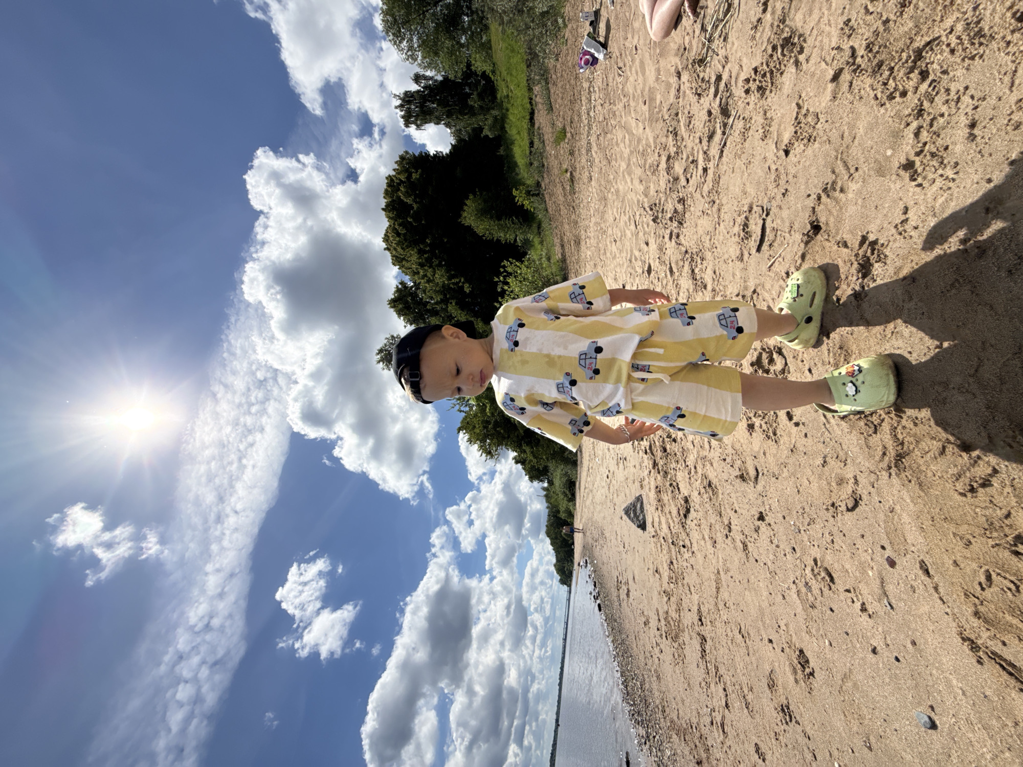 A young child stands on a sun-drenched sandy riverbank or beach, captured from a slightly elevated perspective. The child, likely a boy, wears a yellow and white striped short-sleeved top and matching shorts adorned with small blue car patterns. A dark baseball cap is worn backward on his head, and he has light green clogs on his feet. He is looking downwards or slightly towards the left side of the frame.
The setting is an expansive outdoor area, with a wide stretch of golden sand occupying the foreground and midground. To the left, a calm body of water, presumably a river given the Rusanovo, Russia context, meets the sand. The water surface reflects the bright sky. Along the far bank, a dense line of lush green trees and foliage provides a natural backdrop.
The sky is a vibrant blue, filled with scattered white, fluffy cumulus clouds. The sun shines brightly from the upper left portion of the sky, creating a strong lens flare and casting distinct shadows of the child and other objects onto the sand, indicating it is a sunny day, likely mid-day or afternoon. Footprints and small natural debris are visible on the sand. A bare leg or foot of another person can be seen in the upper right background, suggesting companionship or other people nearby. The overall atmosphere is bright, warm, and peaceful, typical of a pleasant outdoor excursion. No visible text is discernable in the image.