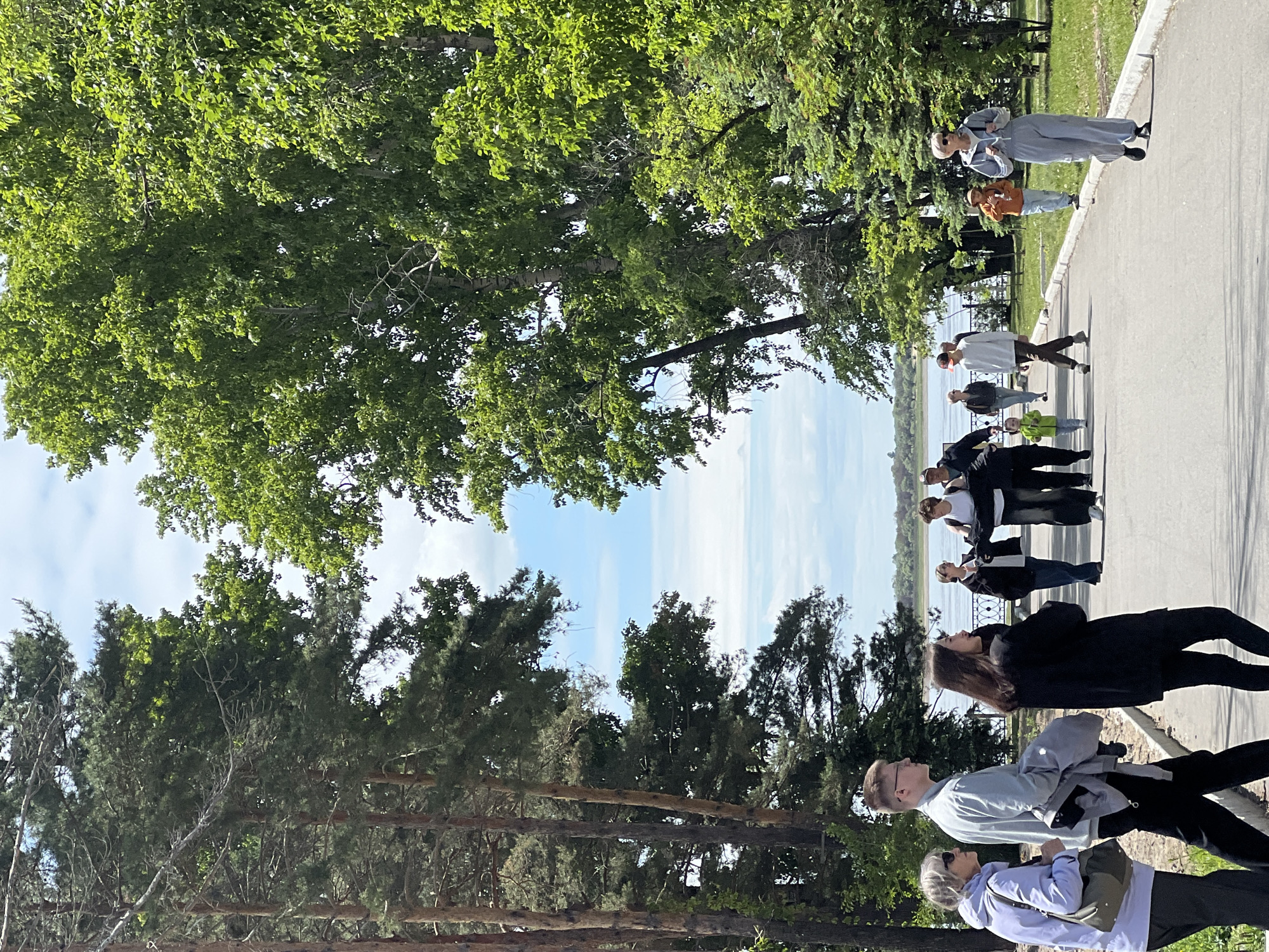 This image depicts a daytime scene along a paved promenade or park path in Khabarovsk, Russia, likely near the Amur River embankment. The sky above is light blue with scattered white clouds.
In the foreground, on the left, three individuals walk away from the camera. The leftmost person is an older woman in a light purple hooded jacket, dark pants, and sunglasses, carrying a dark olive-green shoulder bag. To her right, a young man wears a light blue long-sleeved shirt, dark pants, and glasses, with a grey jacket draped over his left arm. Next to him, a young woman in a black jacket and dark pants looks towards the right.
Further down the path, a diverse group of people is distributed. On the left side of the path, a man in a black top and dark pants walks, accompanied by a young child in a bright green top and light blue jeans. Towards the center of the path, a small group of adults interacts; one woman in a white top and dark pants appears to be reaching towards or interacting with another woman in a black top and dark pants, while a man in a dark top and cap stands behind them. On the right side of the path, a woman in a light grey tracksuit or jumpsuit with dark shoes and sunglasses walks towards the right, carrying a black backpack. To her left and slightly behind, a young boy in an orange top, light pants, and a hat stands near the edge of the path.
The path itself is asphalt and is bordered on the right by a white concrete curb and a grassy area. Beyond the path, on the right, dense green foliage from deciduous and coniferous trees fills the midground. On the left side of the path, tall pine trees with dark green needles line the walkway. In the background, a wide body of water, consistent with the Amur River, stretches horizontally across the frame, with a distant, forested landmass visible on the opposite bank under the horizon.