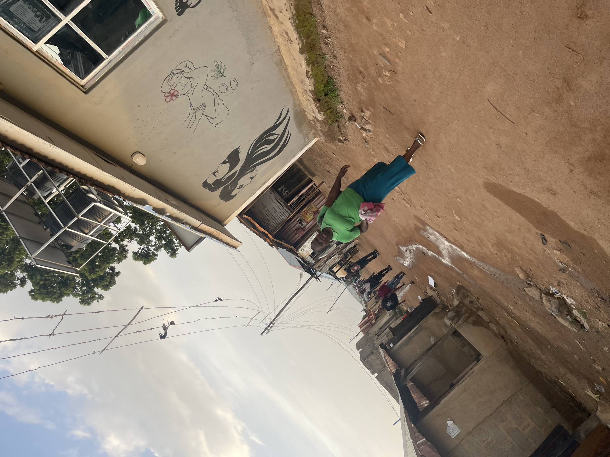 An exterior, low-angle, tilted shot shows a woman walking in a dusty street in Jos, Nigeria. The woman is wearing a bright green t-shirt and a blue skirt. She is carrying a pink and white cloth in her left hand. She is walking away from the camera. The street is unpaved and appears to have puddles of water. On the right side of the street, there is a building with a mural of faces and a woman receiving a massage. There are also a few other people walking in the street in the background. The sky is partly cloudy and the sun is low in the sky. The image appears to have been taken in the late afternoon.