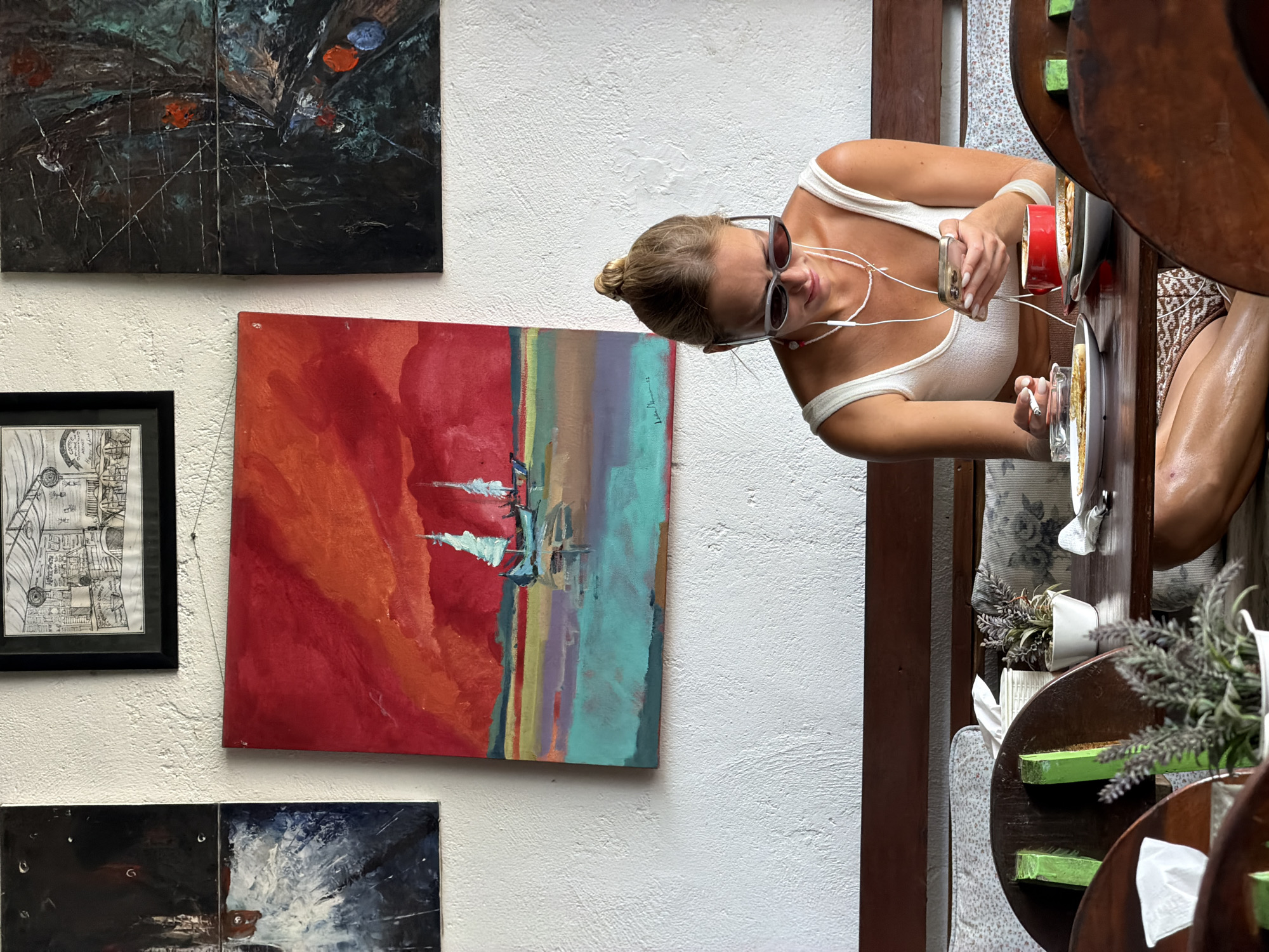 The image captures an indoor scene featuring a young woman seated at a wooden table. She wears a white tank top and sunglasses, has her hair in a bun, and is listening to headphones while holding a cellphone. A lit cigarette rests on a small plate in front of her, next to a half-eaten meal.
Behind her, a wall displays an assortment of artwork. The paintings include an abstract piece featuring shades of red and turquoise and another darker abstract work in shades of black. There is a framed black and white print. The wall is textured with a white finish.
The setting appears to be a cafe or restaurant. Given the request, the location is likely in North Kuta, Indonesia. Other patrons are partially visible in the background. The wooden table is cluttered with items including napkins and potted lavender.