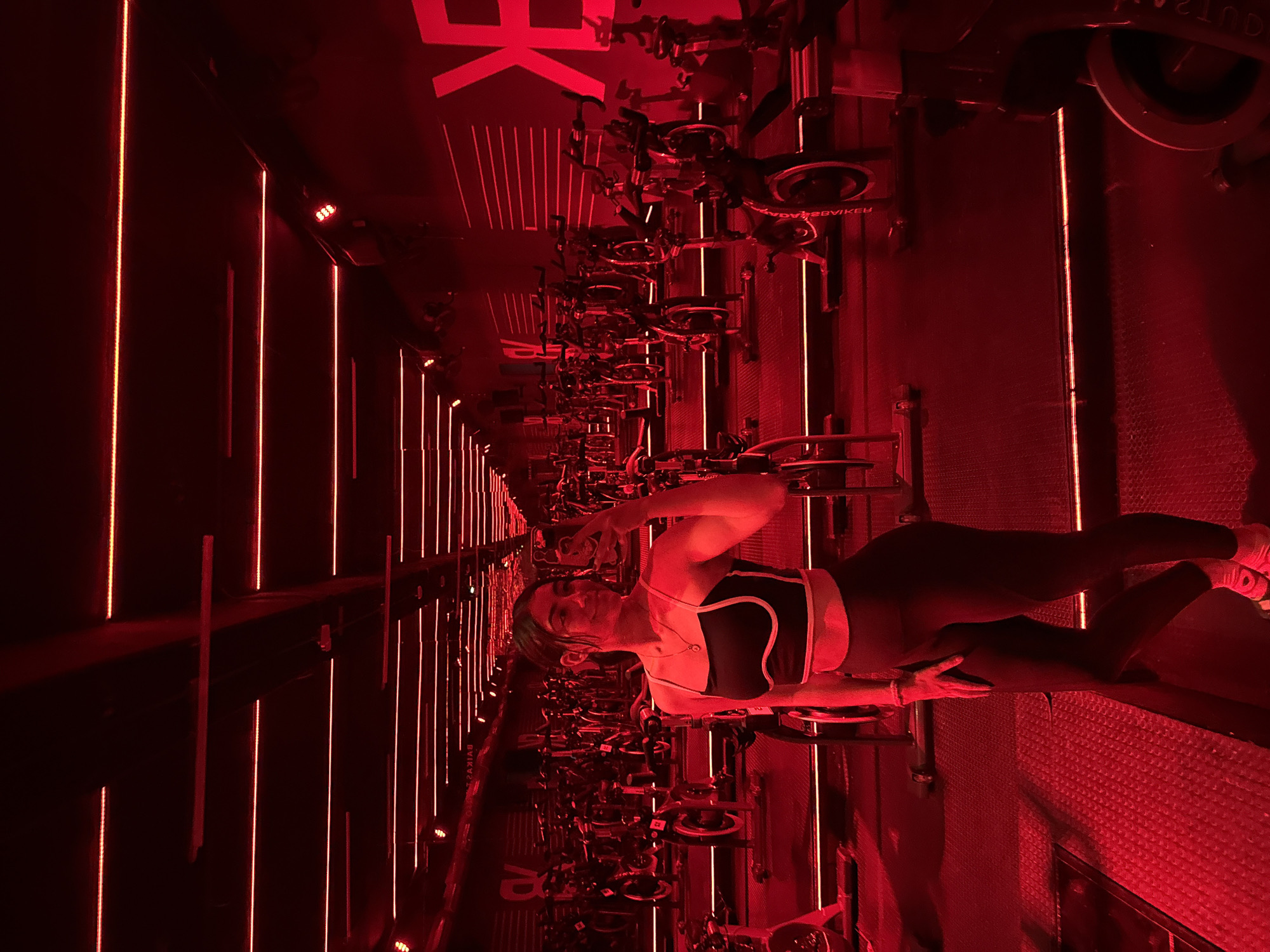 A woman is standing among rows of stationary bikes in a dimly lit room with red lighting, presumably in a fitness studio. The lighting includes vertical red lines on the walls. The woman is wearing dark athletic wear and sneakers. The stationary bikes are predominantly black. The location is implied to be a fitness studio in Puebla, Mexico.