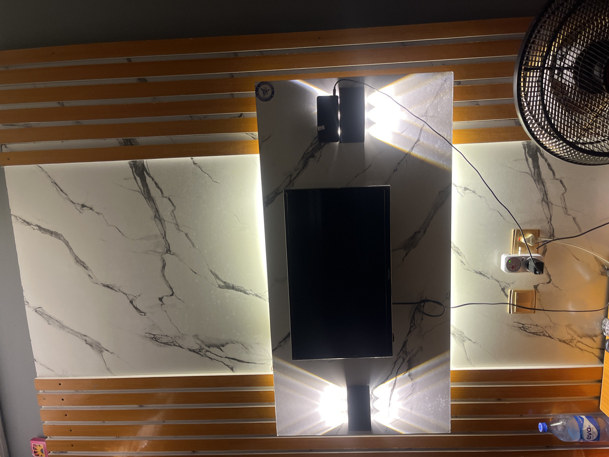 The image depicts an indoor scene, likely a living room or entertainment area, featuring a wall-mounted television. The wall behind the television has a distinctive white marble pattern with black veins, illuminated by hidden strip lighting that casts a warm glow. Flanking the television are vertical wooden slats with an orange hue, creating a modern aesthetic. Two wall-mounted light fixtures, positioned on either side of the television stand, emit upward and downward beams of light, highlighting the textured marble. Below the television, a dark-colored power strip is visible, connected to electrical outlets. To the left, a bottle of Eva water is placed on a surface, alongside what appears to be a small digital device. On the right side of the frame, a black oscillating fan is partially visible. The overall impression is of a contemporary and well-lit interior space. The prompt indicates that this scene is located in Ajara, Nigeria.