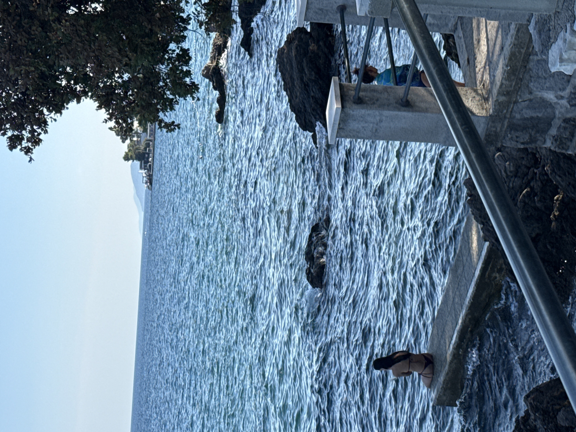The image depicts a coastal scene in Opatija, Croatia, on a clear day with calm, blue waters. In the foreground, a person in a bikini is seated on a concrete platform extending into the sea. To the right, a second person, partially obscured by a metal railing, stands on a paved walkway with a concrete railing. The background shows rocky shores, trees, and distant buildings along the coastline, under a bright sky.