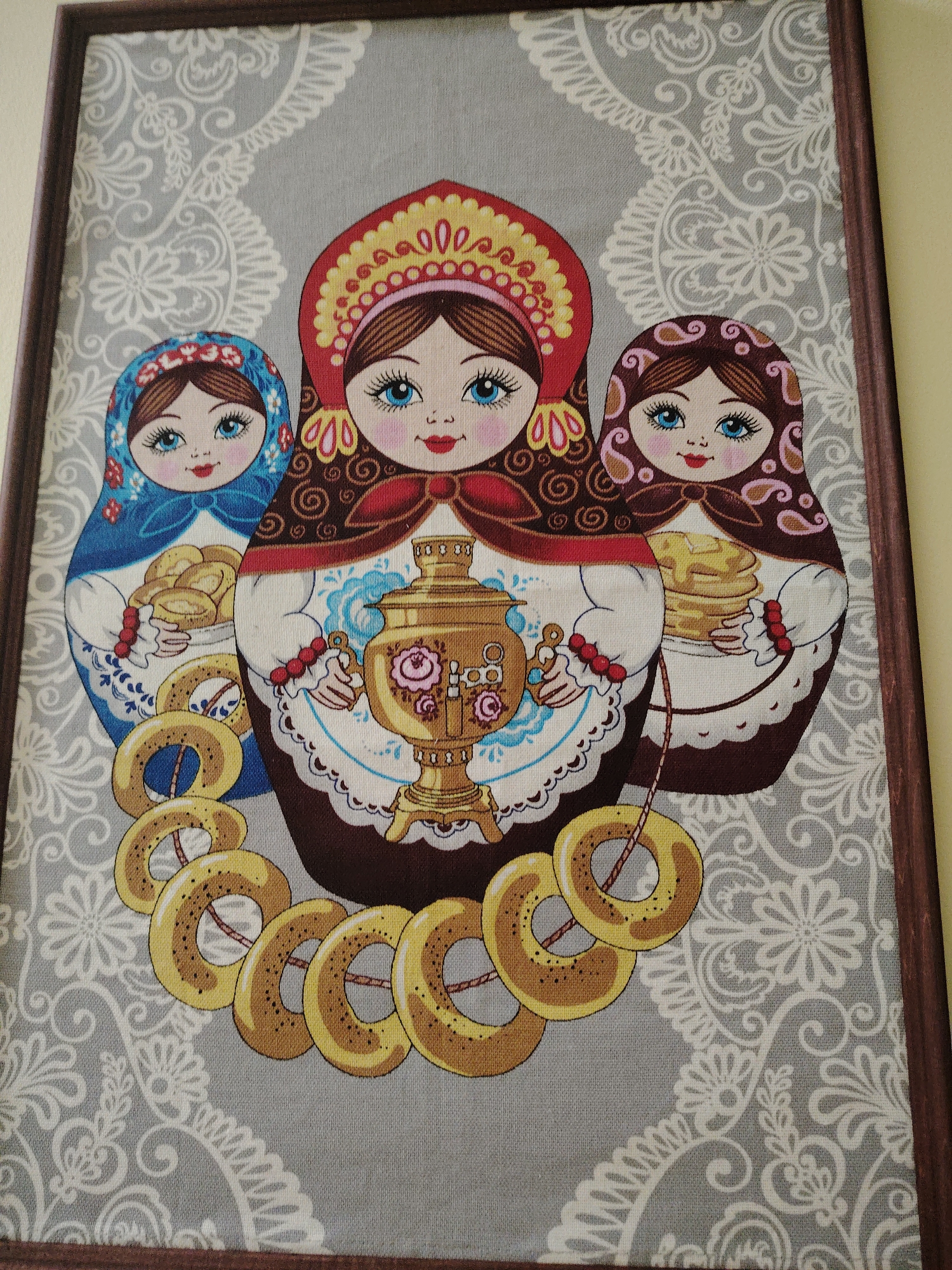 This framed textile artwork depicts three stylized Russian Matryoshka-like figures against a patterned gray background. The central figure is the largest, wearing a red and yellow kokoshnik-style headpiece and a dark brown dress with a red bow. She holds a golden samovar, adorned with pink roses, ready to serve tea. To her left, a smaller doll in a blue headscarf and dress holds a basket filled with what appear to be small buns or pirozhki. To the central figure's right, another smaller doll in a brown headscarf with pink patterns and a brown dress holds a stack of blini or pancakes.
All three dolls have fair skin, prominent blue eyes, rosy cheeks, and smiling red lips, conveying a cheerful and welcoming expression. A garland of golden, ring-shaped bread, traditionally known as sushki or baranki, is draped across the lower portion of the image, connecting the figures.
The background is a soft gray fabric, patterned with intricate white floral and swirling designs that resemble traditional lacework or brocade. The entire scene is enclosed within a simple dark brown wooden frame. The artwork celebrates elements of Russian culture and hospitality, suggesting a festive gathering or tea ceremony. No visible text is present in the image. The media file is a photograph of this framed artwork, likely depicting a cultural piece from Russia, aligning with the provided location context of Sarapul, Russia.