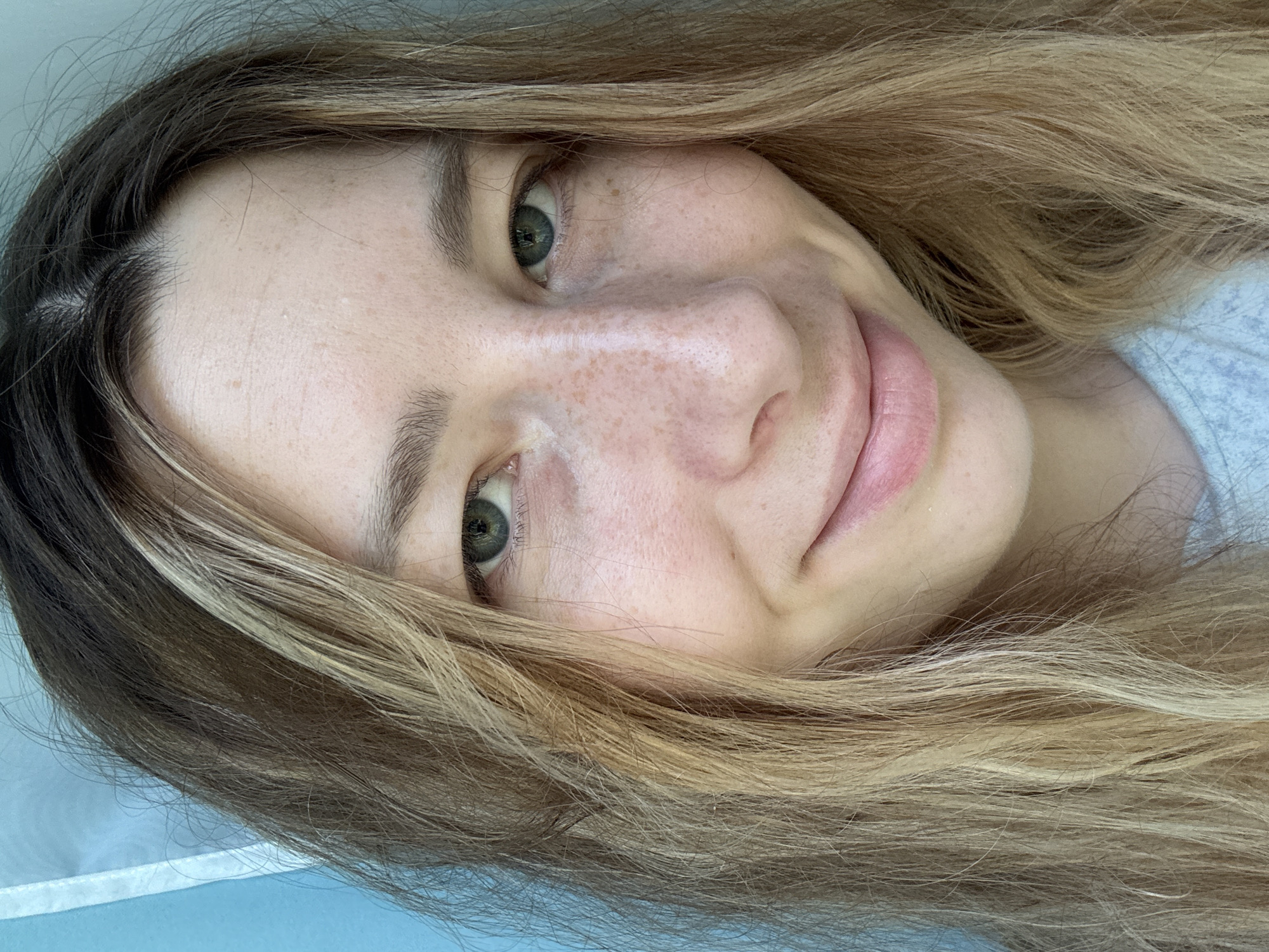 A close-up, angled view of a Caucasian woman with fair skin and freckles. Her hair is long, wavy, and appears to be dyed with dark brown roots and blonde highlights. The woman has green eyes and is smiling slightly. The background appears to be a soft, blurred, light blue. The location in Moscow, Russia is not determinable.