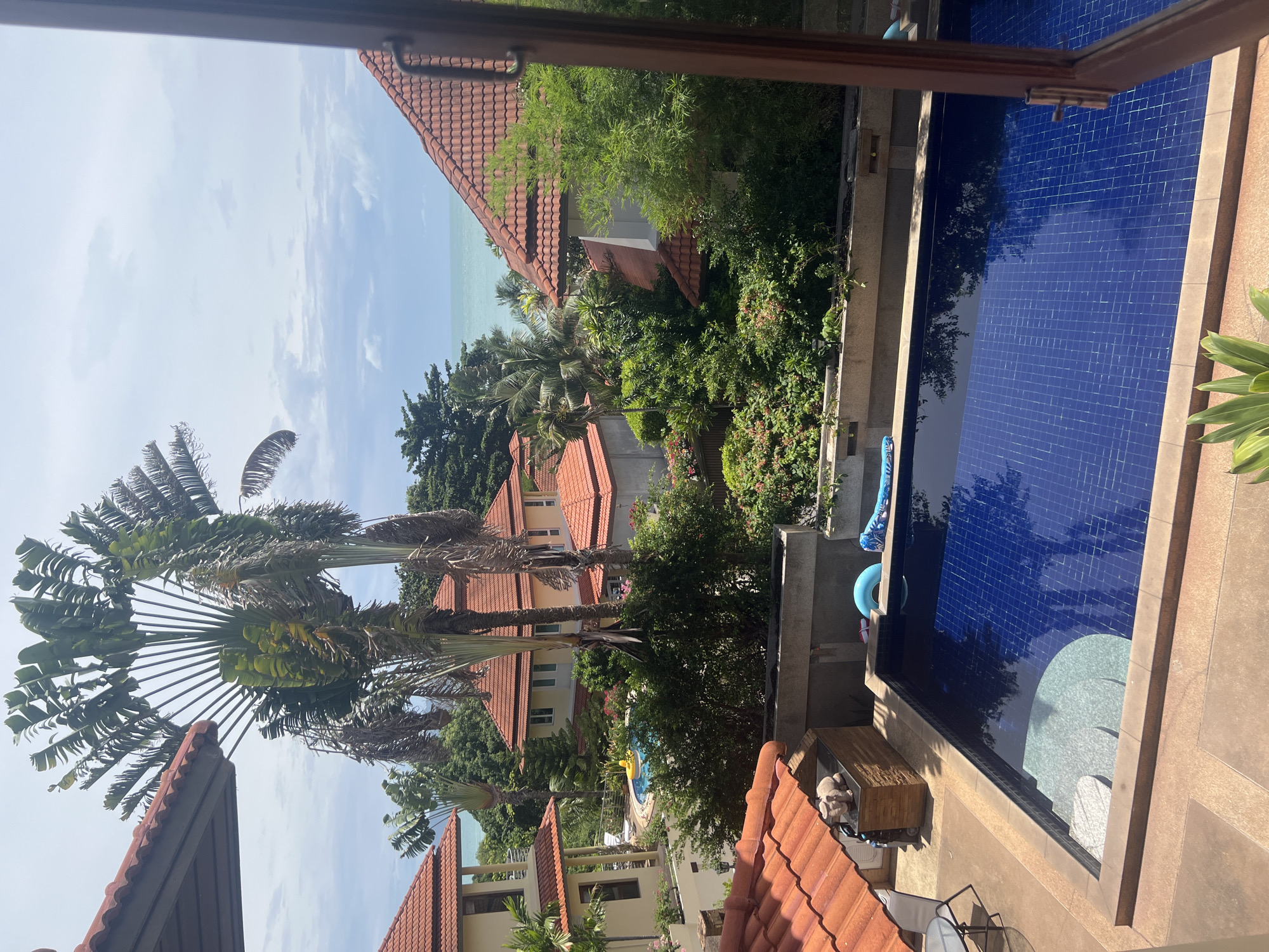 This is an outdoor, eye-level, medium shot, captured on a sunny day in Ko Samui District, Thailand. The scene features a swimming pool with bright blue tiles, surrounded by tan-colored paving. Steps with light gray textured surfaces lead into the pool. On the edge of the pool, a blue inflatable ring and a deflated blue float are visible. To the left of the pool, there is an outdoor grill area with a wooden and stone counter, topped with a light-colored elephant figurine. A table and chairs are also present on the patio.
In the background, lush green vegetation, including palm trees and flowering bushes, creates a tropical ambiance. Several houses with red tiled roofs are scattered amongst the greenery. Beyond the houses and trees, the turquoise ocean stretches to the horizon under a partly cloudy sky. The image is taken from a vantage point that includes part of a wooden window frame on the right side, suggesting it was taken from inside a room or covered area looking outwards. There are no people visible in the scene.