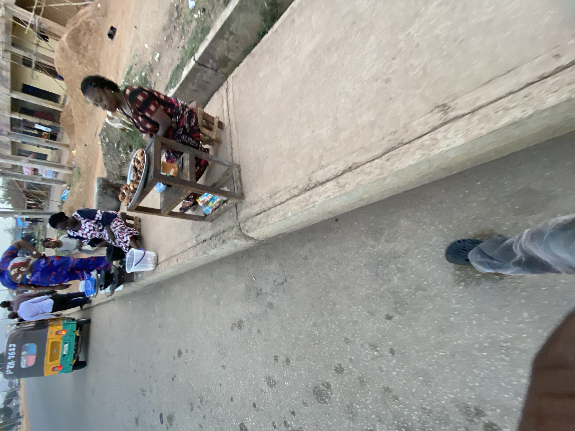 The image captures a bustling roadside scene in Bukuru, Nigeria, from a slightly elevated perspective. In the foreground, the photographer's left foot and leg are partially visible, suggesting a personal viewpoint.
The main subjects are several women engaged in street commerce along a concrete sidewalk. Centrally, a woman in a patterned red and black dress stands behind a simple metal stand, attending to a tray of food items, possibly fried snacks, displayed for sale. To her left, another woman in a patterned blue and white dress is bent over near a white plastic bucket, while another woman is visible sitting nearby. Further down the street, a man in a white shirt stands near a yellow and green auto-rickshaw (often called a Keke Napep), which has "PTA-1613" clearly visible on its rear.
The setting includes a paved asphalt road, a raised concrete sidewalk, and rough terrain with some buildings and sparse vegetation in the background. The buildings appear to be simple structures, some possibly unfinished or older. Plastic buckets are scattered near the vendors.
The overall activity depicts daily life and informal street vending. The scene appears to be taking place during the daytime under clear weather conditions, indicated by the bright, natural light and lack of heavy shadows. There are no strong emotional expressions visible, with individuals focused on their tasks or interactions.